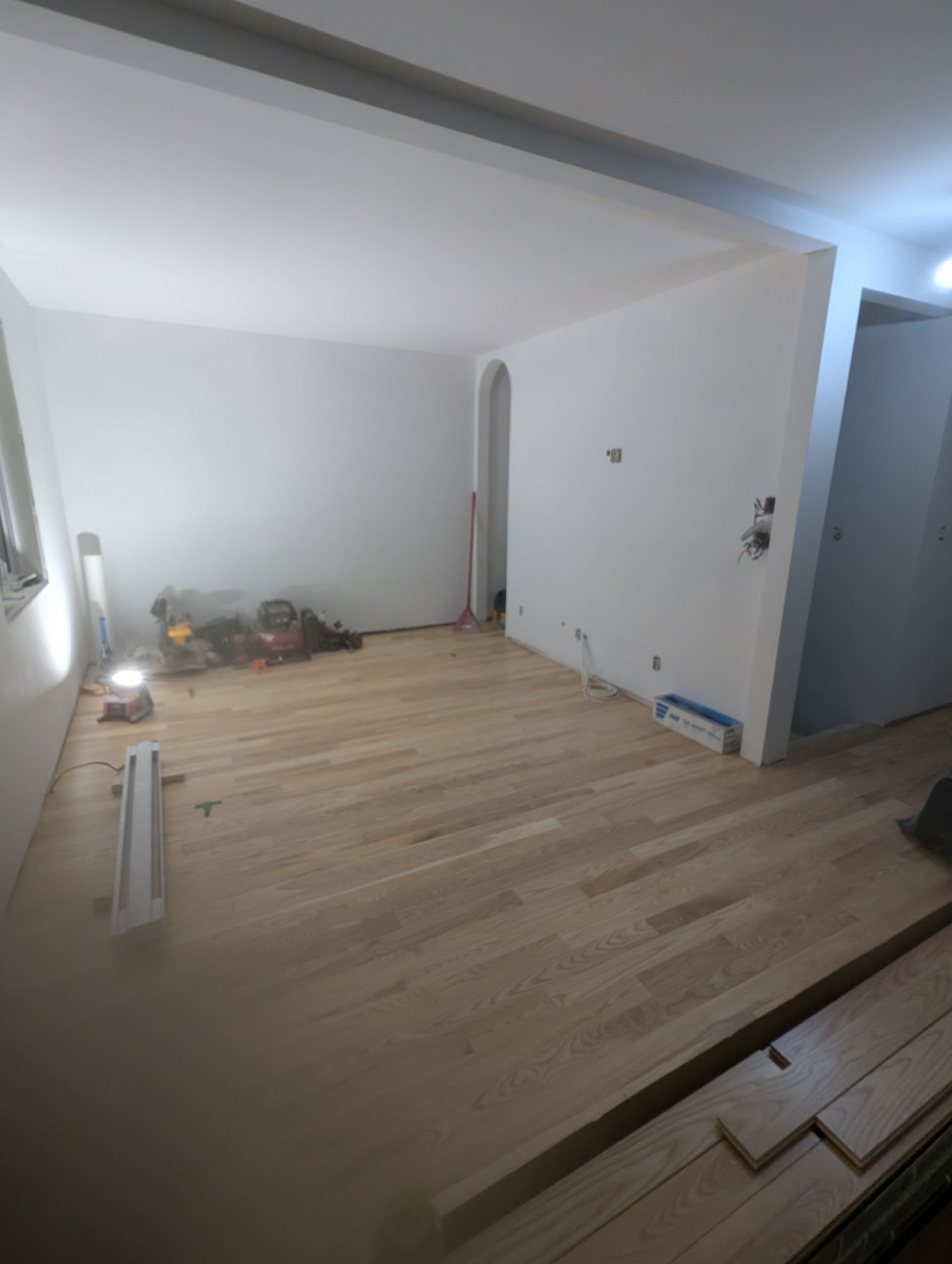 Pièce vide avec parquet neuf, murs blancs et outils éparpillés, probablement en cours de rénovation.
