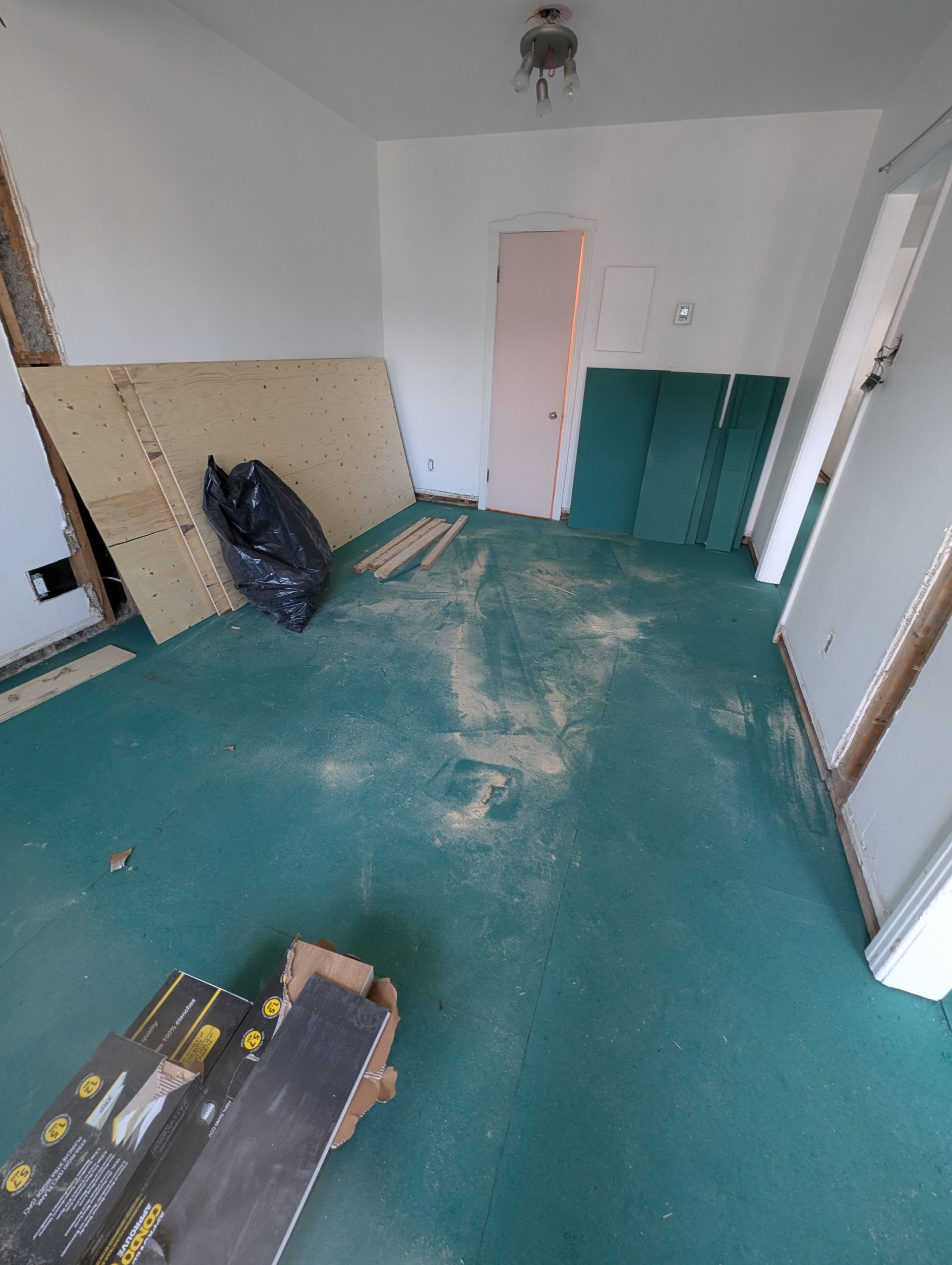 Pièce en travaux avec sol turquoise, contreplaqué et débris. Sol stratifié et porte rose.