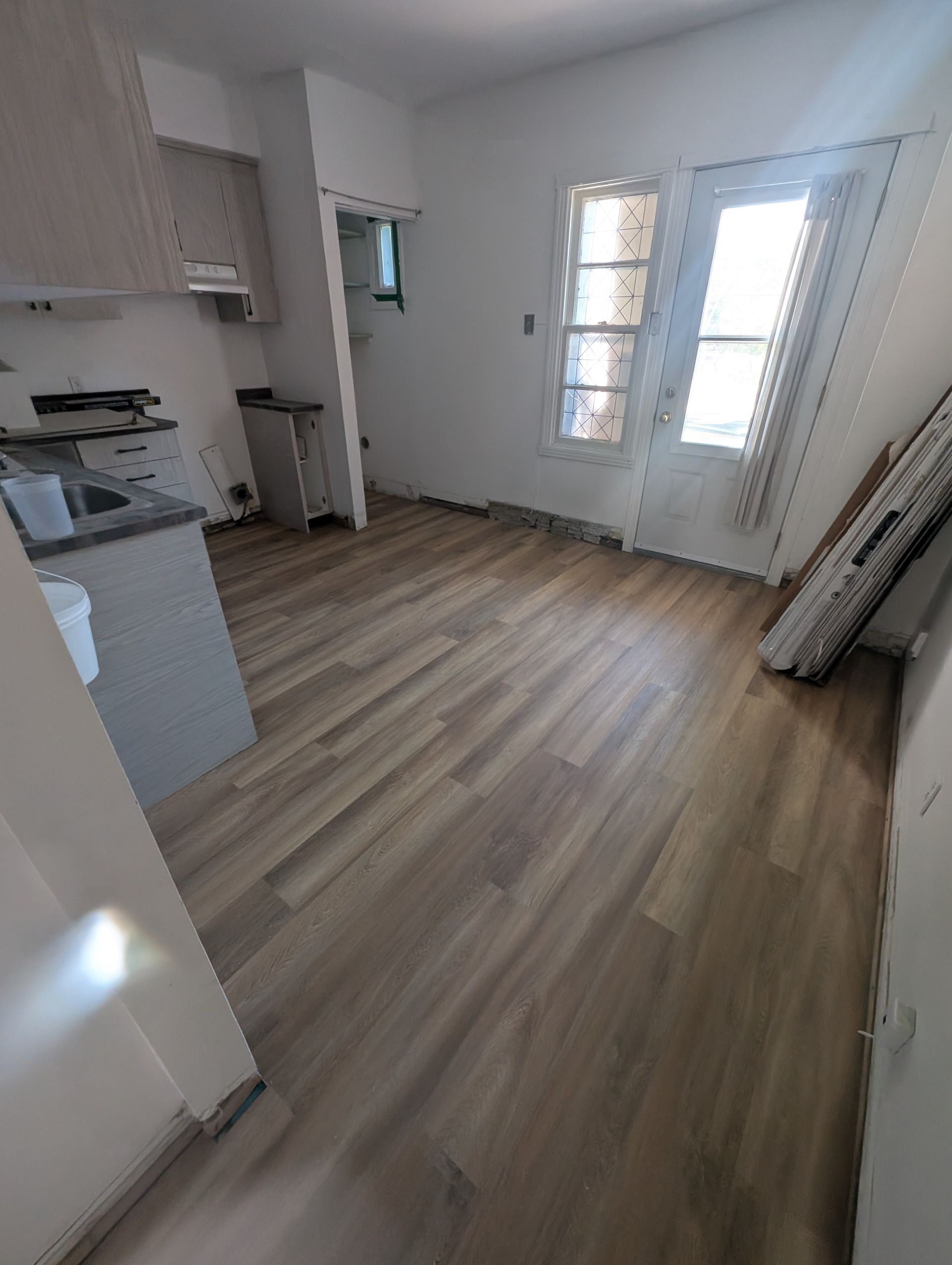 Intérieur d'appartement vide avec parquet clair, une cuisine et une porte donnant sur une fenêtre lumineuse.
