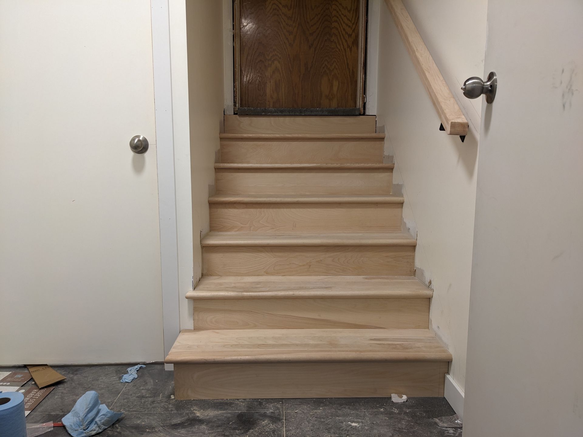 Escalier en bois menant à une porte. Rambarde à droite. Murs beiges, porte fermée à gauche.
