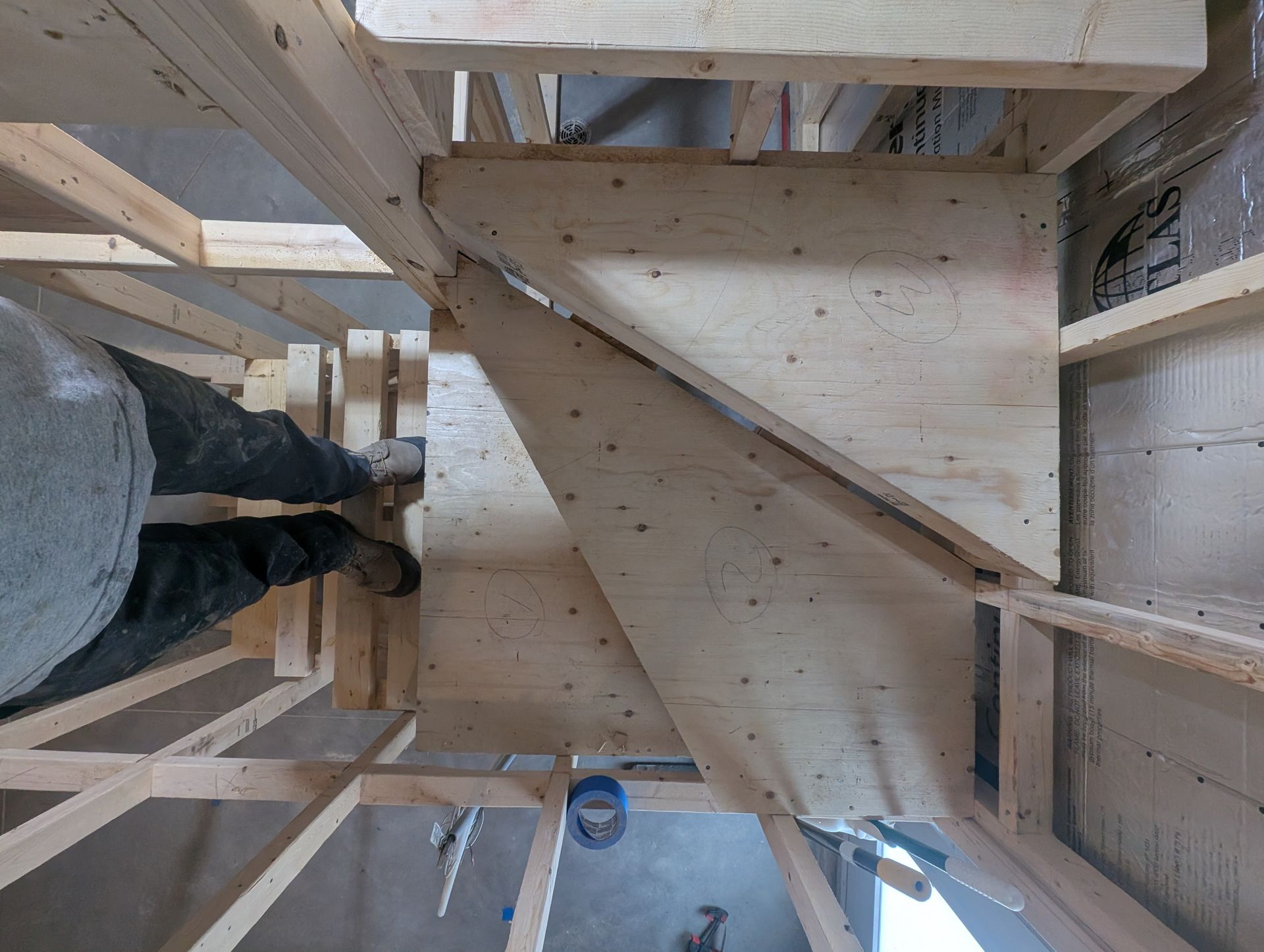 Construction d'un escalier en bois, vue de dessus. Pieds d'un homme posés sur un palier en contreplaqué.