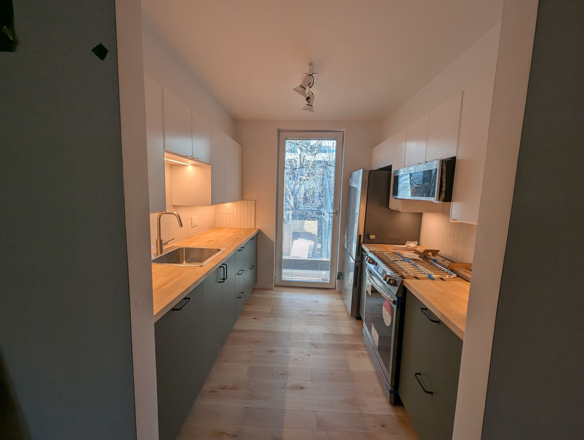 Cuisine longue et étroite avec plans de travail en bois, placards gris et une porte au fond.