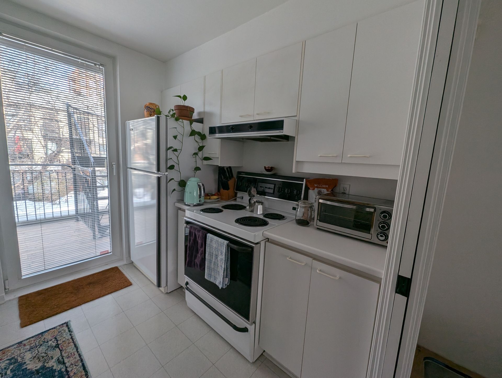 Cuisine blanche équipée d'appareils électroménagers, dont une cuisinière, un réfrigérateur et un micro-ondes, et d'une porte donnant sur un balcon.