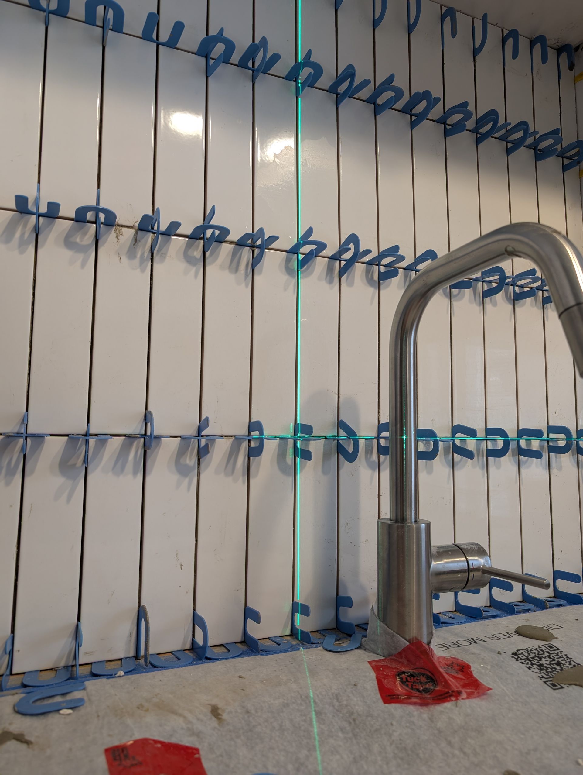 Pose de carrelage sur un mur de cuisine, l'alignement étant vérifié à l'aide d'un niveau laser. Un robinet est visible au premier plan.