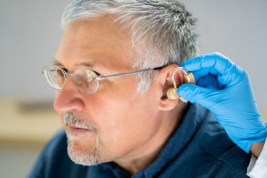 Gloved hand fitting a hearing aid into an older man’s ear
