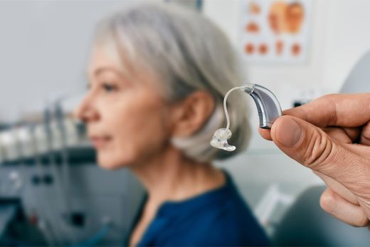Hearing aid held in foreground with older woman blurred in a clinic background