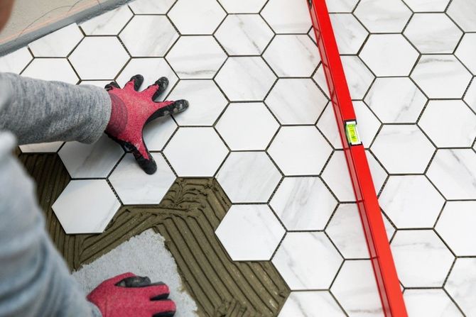Hands in pink gloves laying hexagonal white tiles beside red spacer line on a floor.