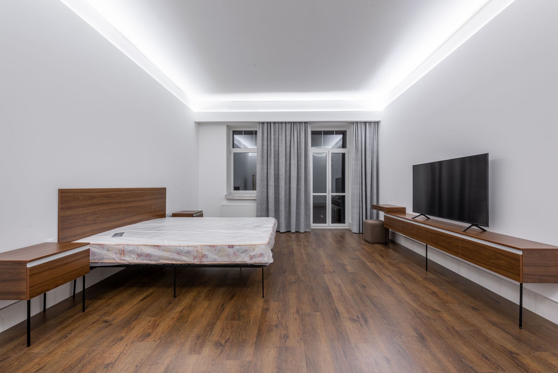 Modern minimalist bedroom with bed, wood floor, curtains, and TV console under bright ceiling lights