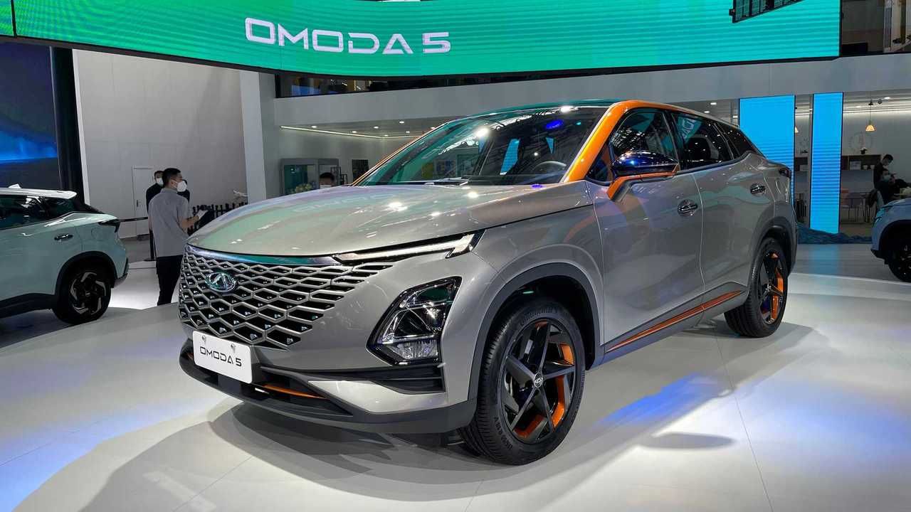 Silver Omoda 5 SUV on display at an auto show, with orange accents and a white car in the background