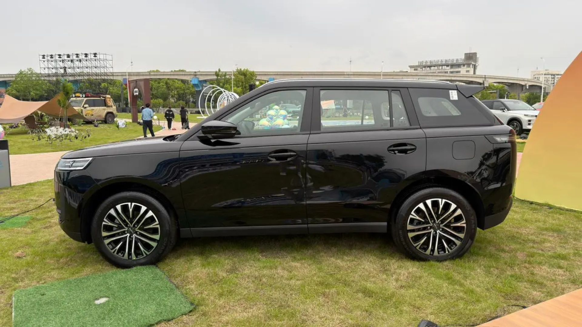 Black SUV parked on grass at an outdoor event, side view with people and buildings in the background