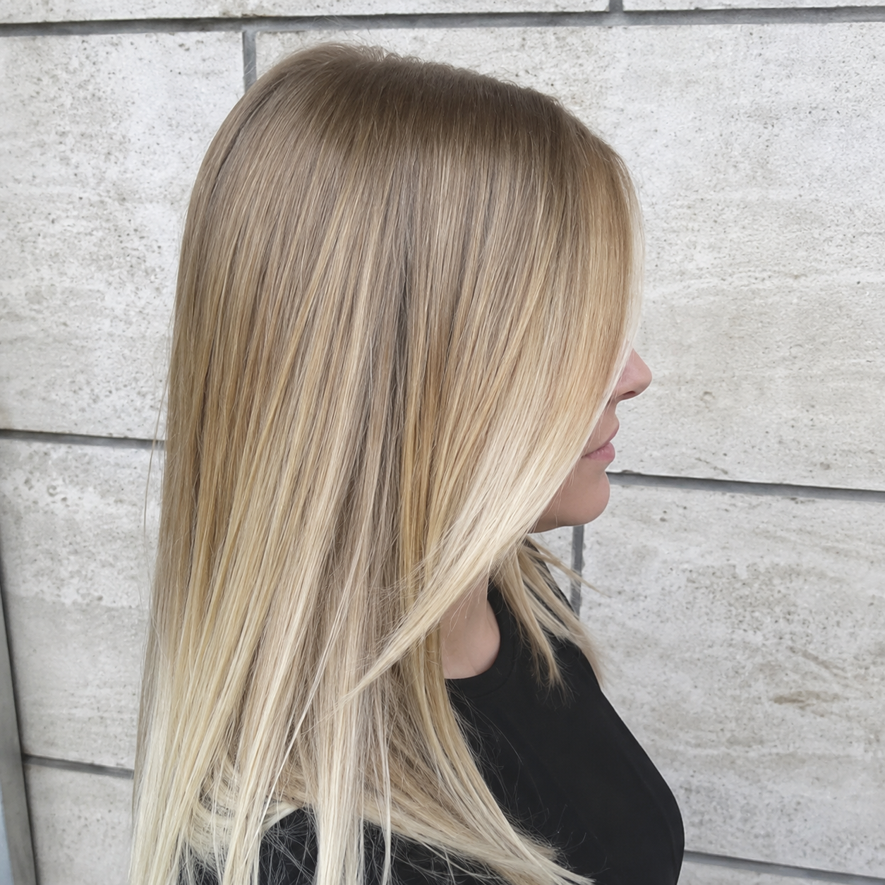 Profilo di una donna bionda con i capelli lisci contro un muro di mattoni grigio chiaro.