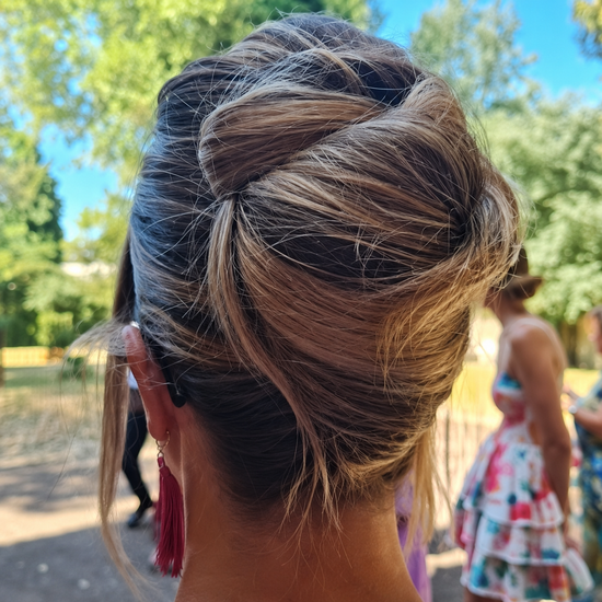 Primo piano di un'elegante acconciatura raccolta intrecciata con riflessi biondi, scattata all'aperto, con una persona sfocata sullo sfondo.