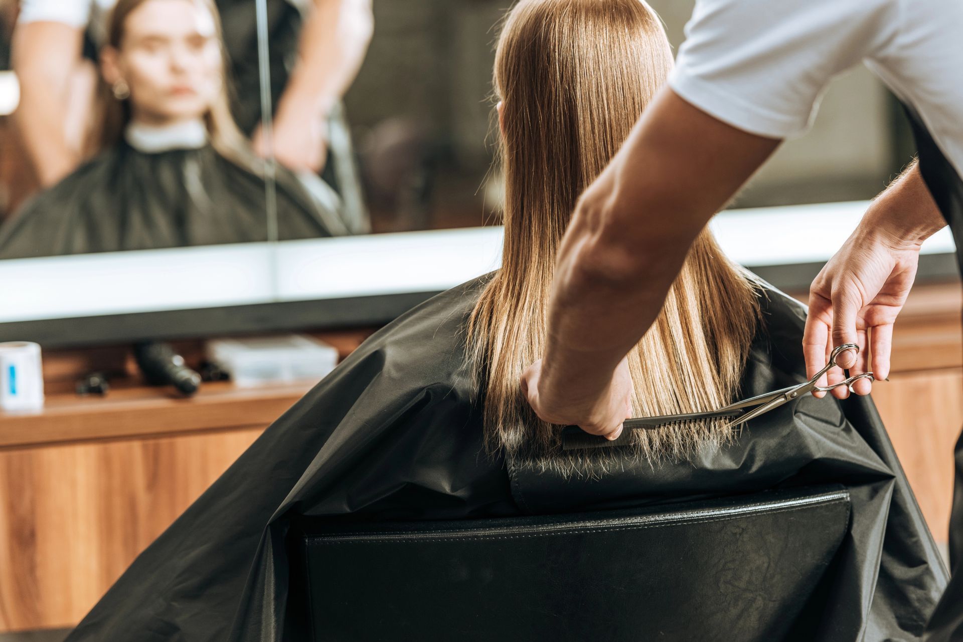 In un salone, una parrucchiera taglia i lunghi capelli di una cliente, con uno specchio che riflette la scena.