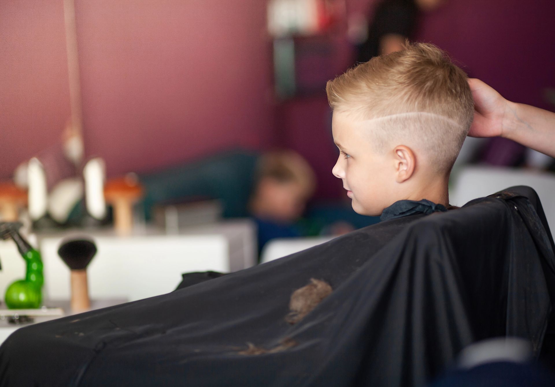 Un bambino si fa tagliare i capelli in una barberia, con un mantello nero e una mano che gli sistema i capelli.