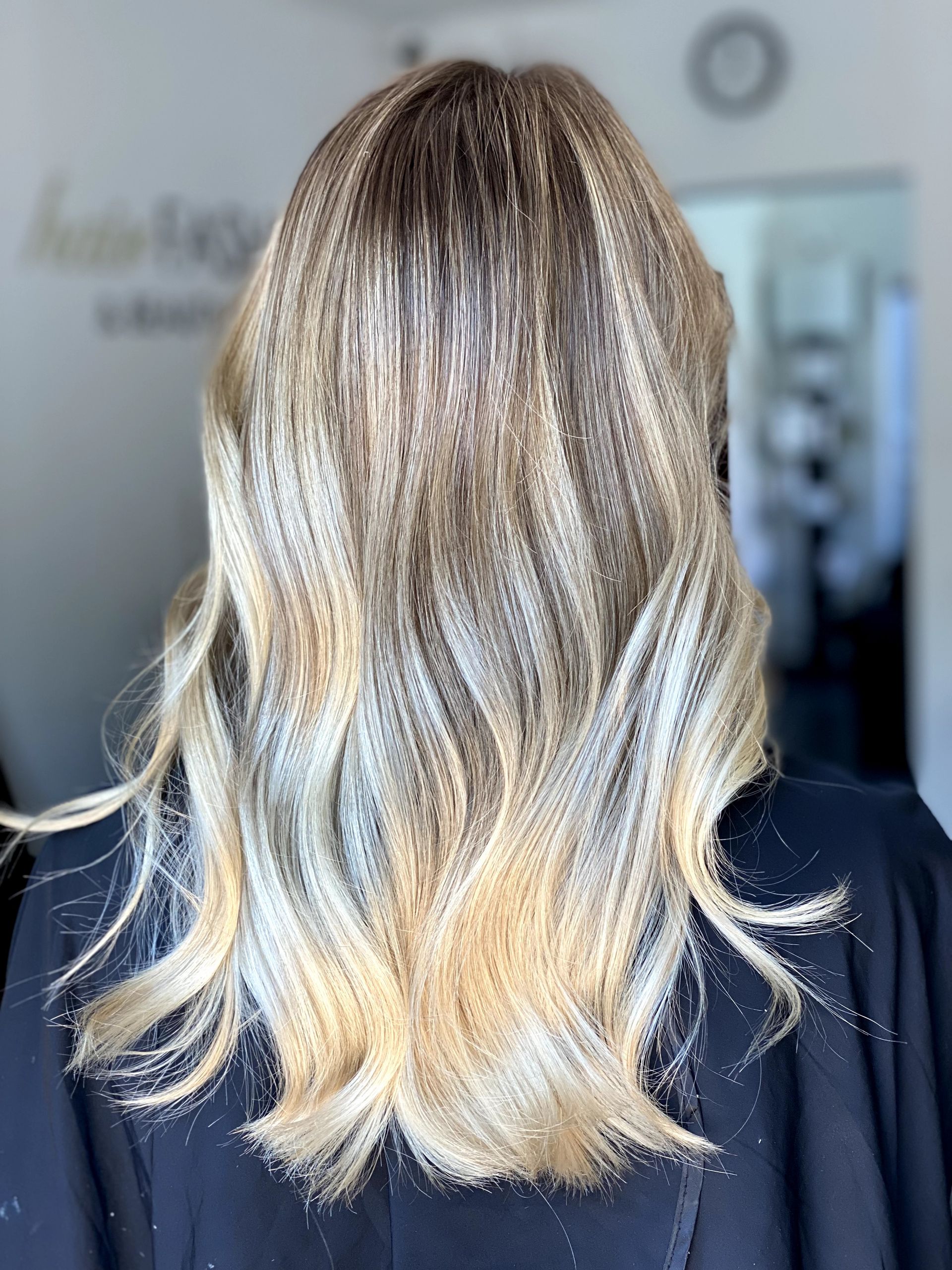 Capelli biondi, mossi, lunghi fino alle spalle con radici più scure, su uno sfondo sfocato di un salone di bellezza.