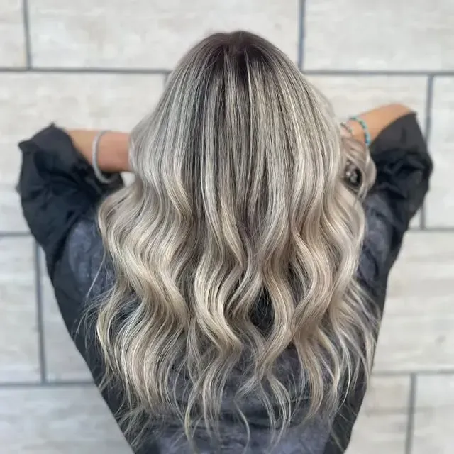 Lunghi capelli mossi color biondo platino con radici scure, visti di spalle contro una parete piastrellata.