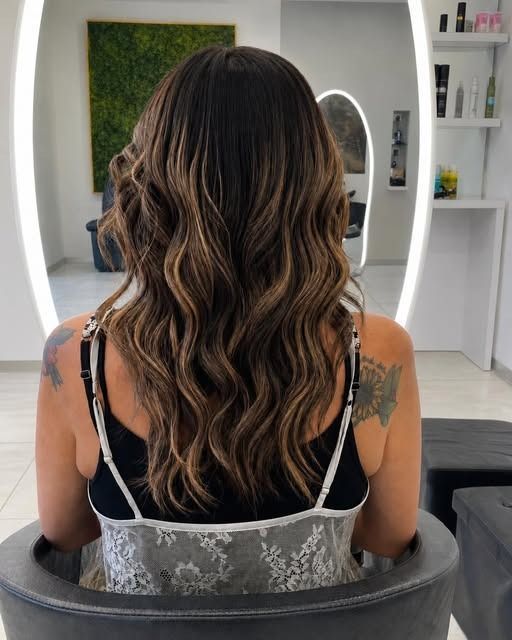 Persona con giacca nera e capelli castani ondulati, vista di spalle contro un muro di pietra.