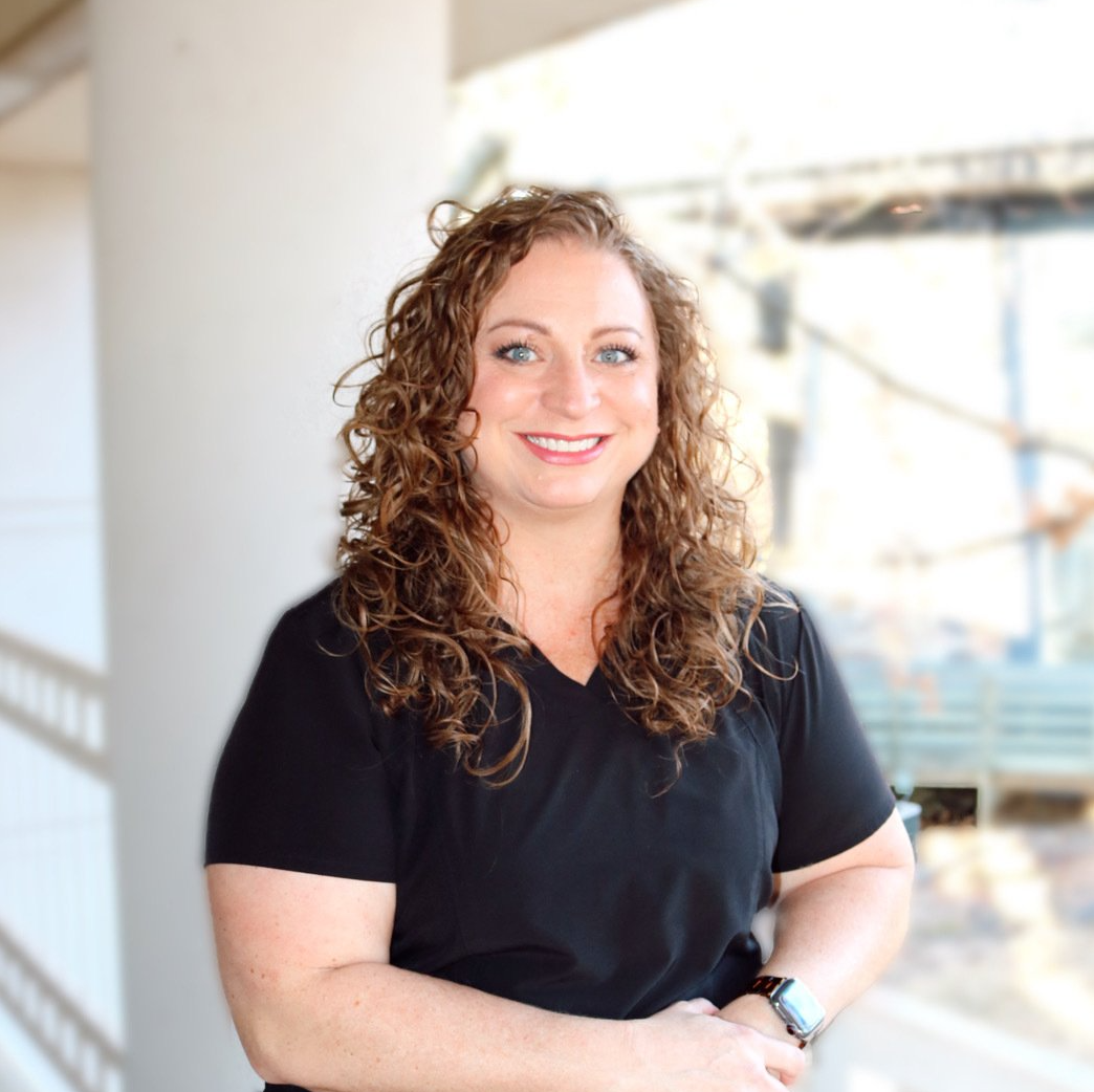 A woman with curly hair is wearing a black scrub top and smiling for the camera.