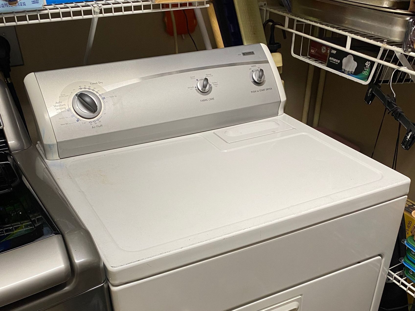 A white Whirlpool dryer with control knobs on top, placed in a laundry area.