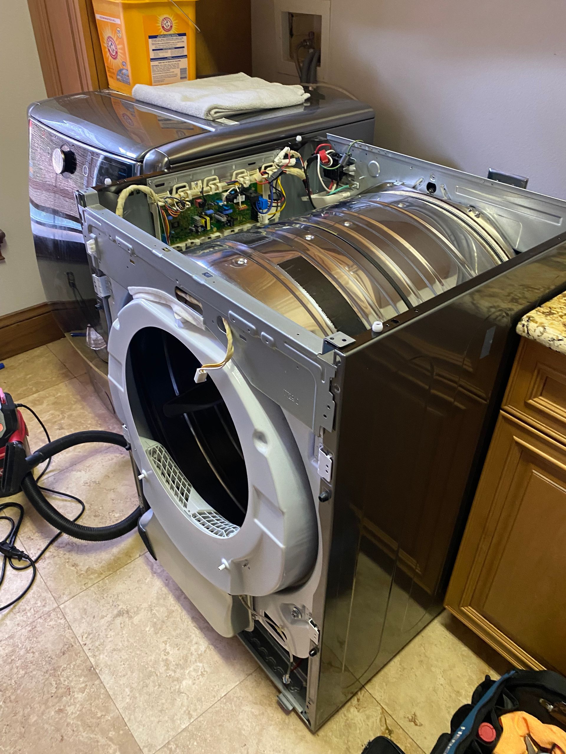 A disassembled silver and black dryer in a laundry room, revealing internal components.