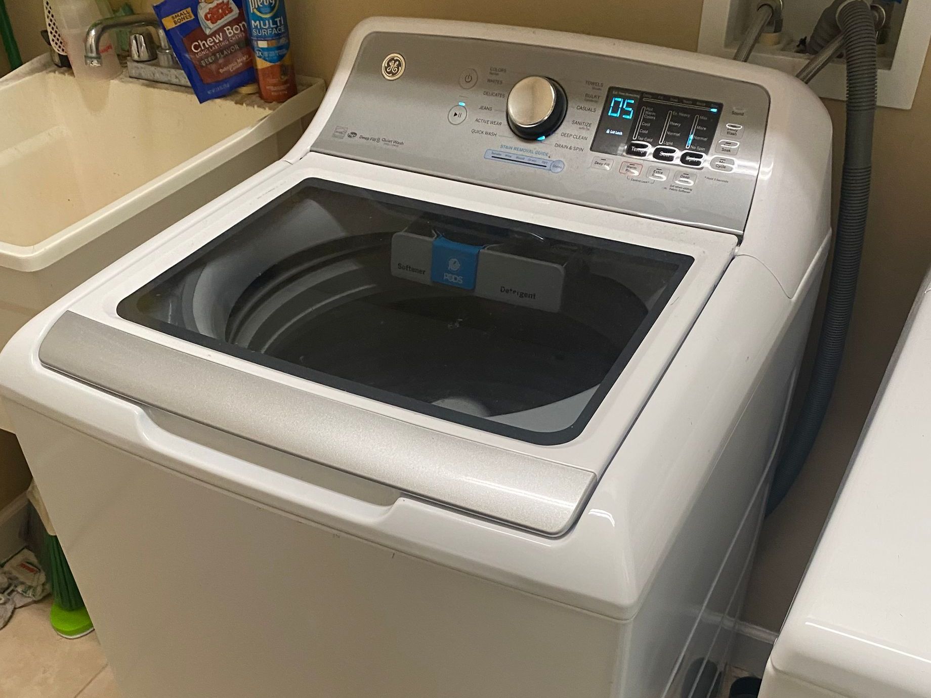 White GE top-load washing machine with control panel and open glass lid next to a utility sink.
