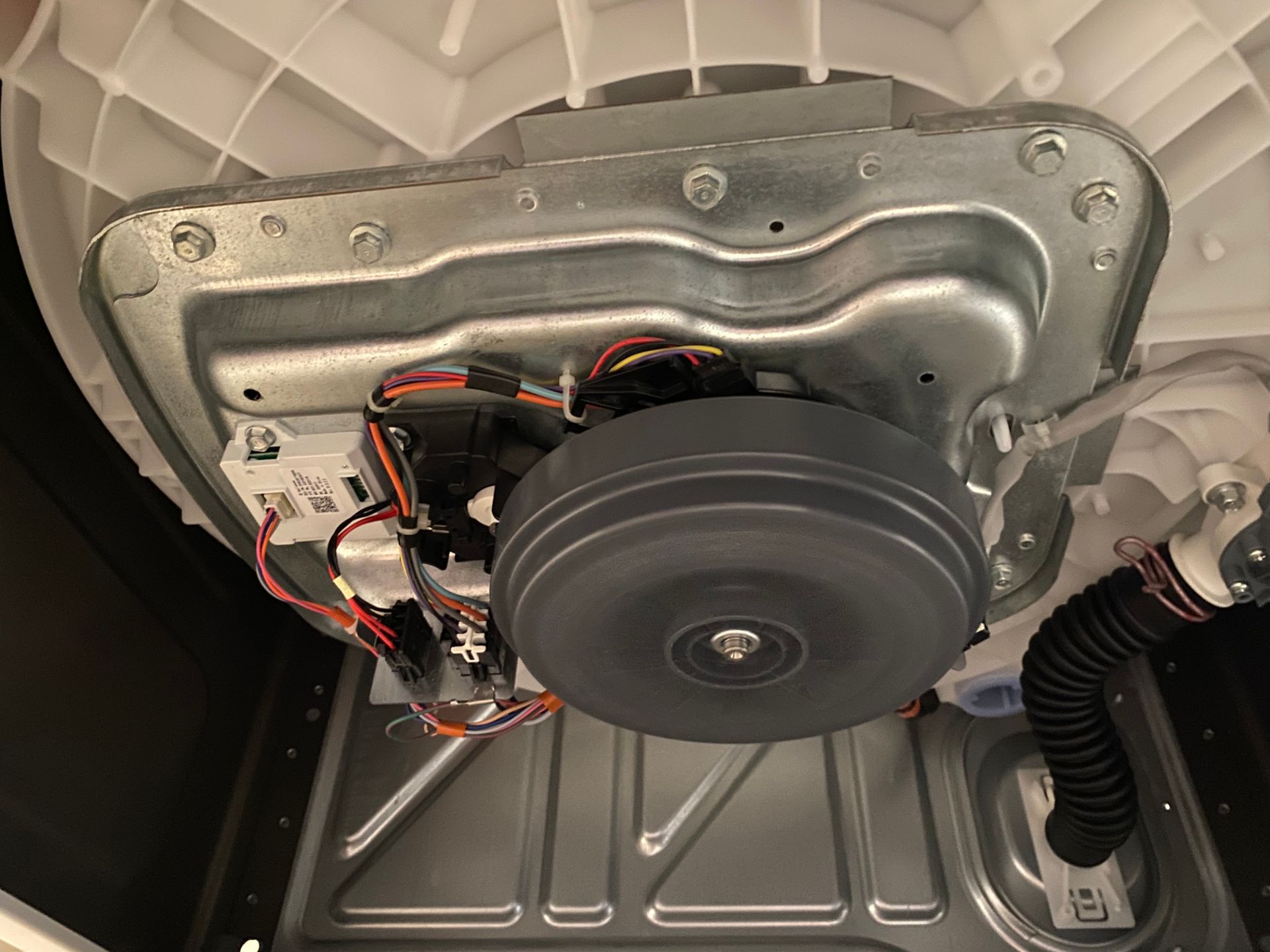 Underside of a washing machine. A large grey disc-shaped motor is visible, with wiring and metal components.