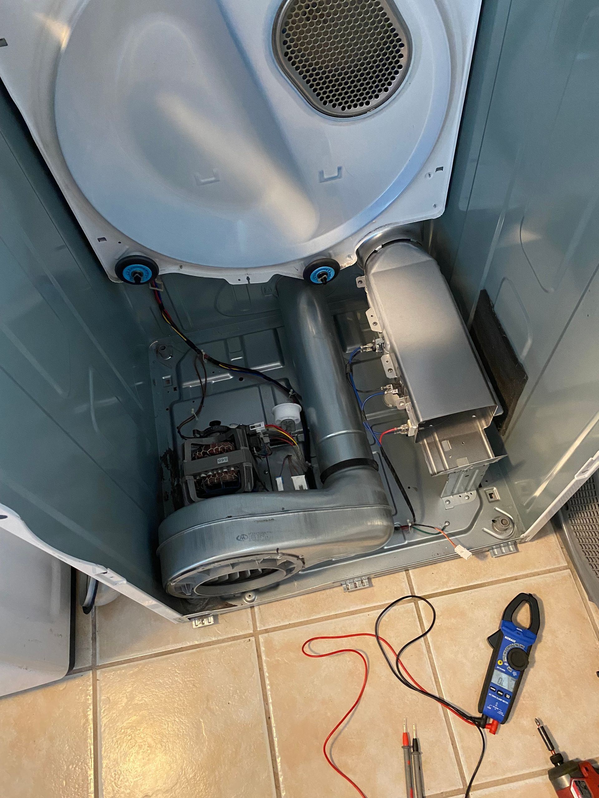 Interior view of a dryer with the back panel removed, showing components like the blower and motor, and a multimeter.