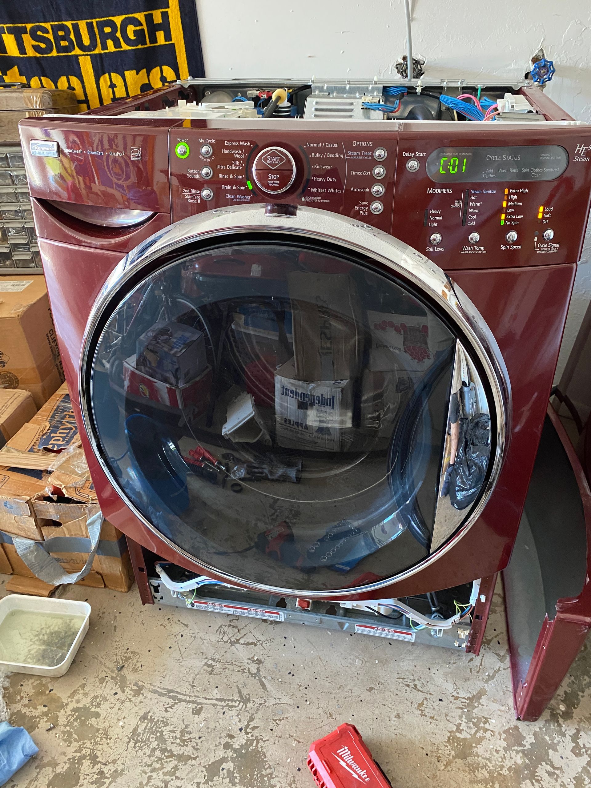 A burgundy front-load washing machine with its top panel removed, displaying internal components.