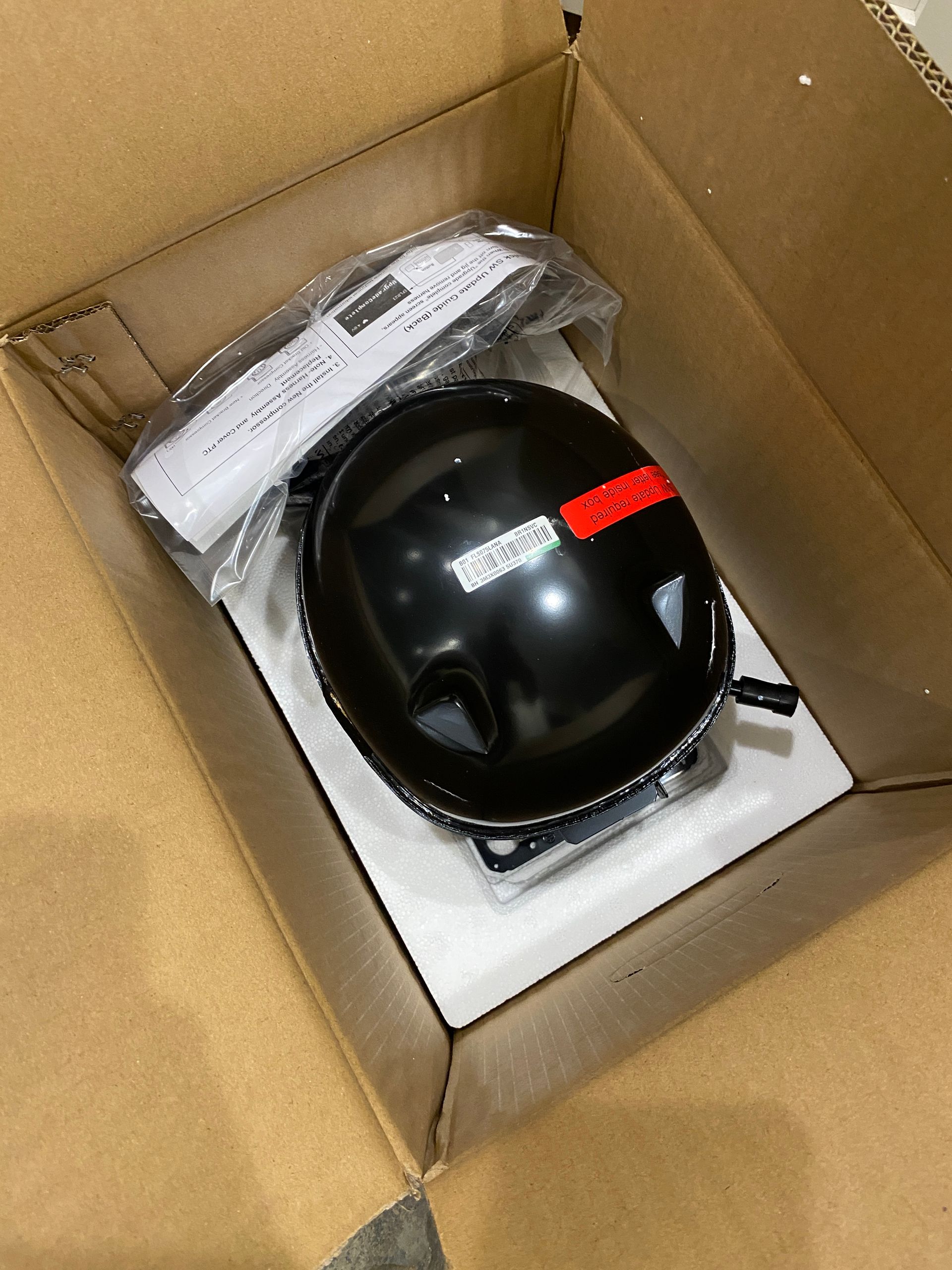 Black refrigerator compressor in a cardboard box, with paperwork.
