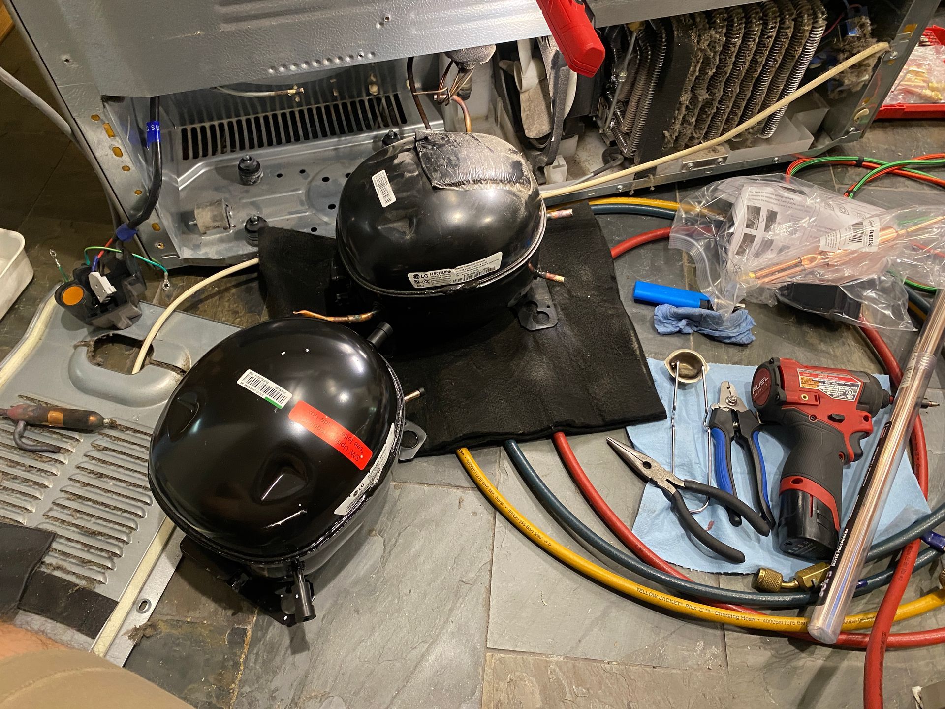 Two black refrigerator compressors and tools on the floor near the refrigerator's back.