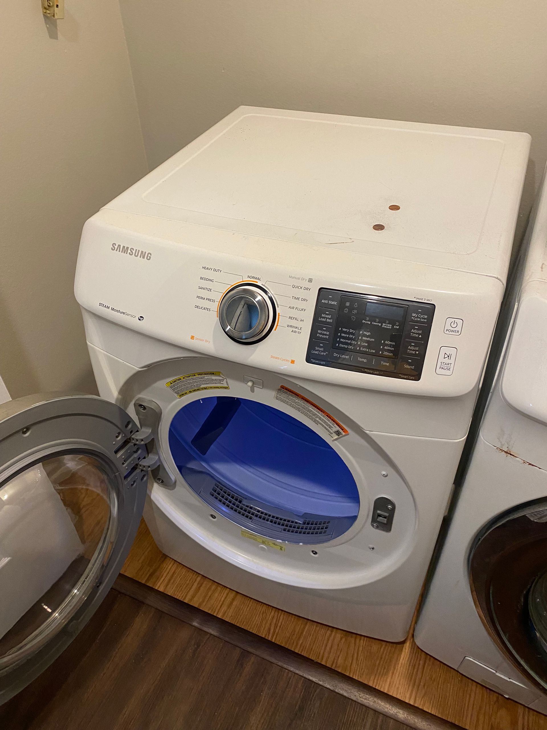 White Samsung dryer with an open door, blue interior, set on a wood floor in a room.