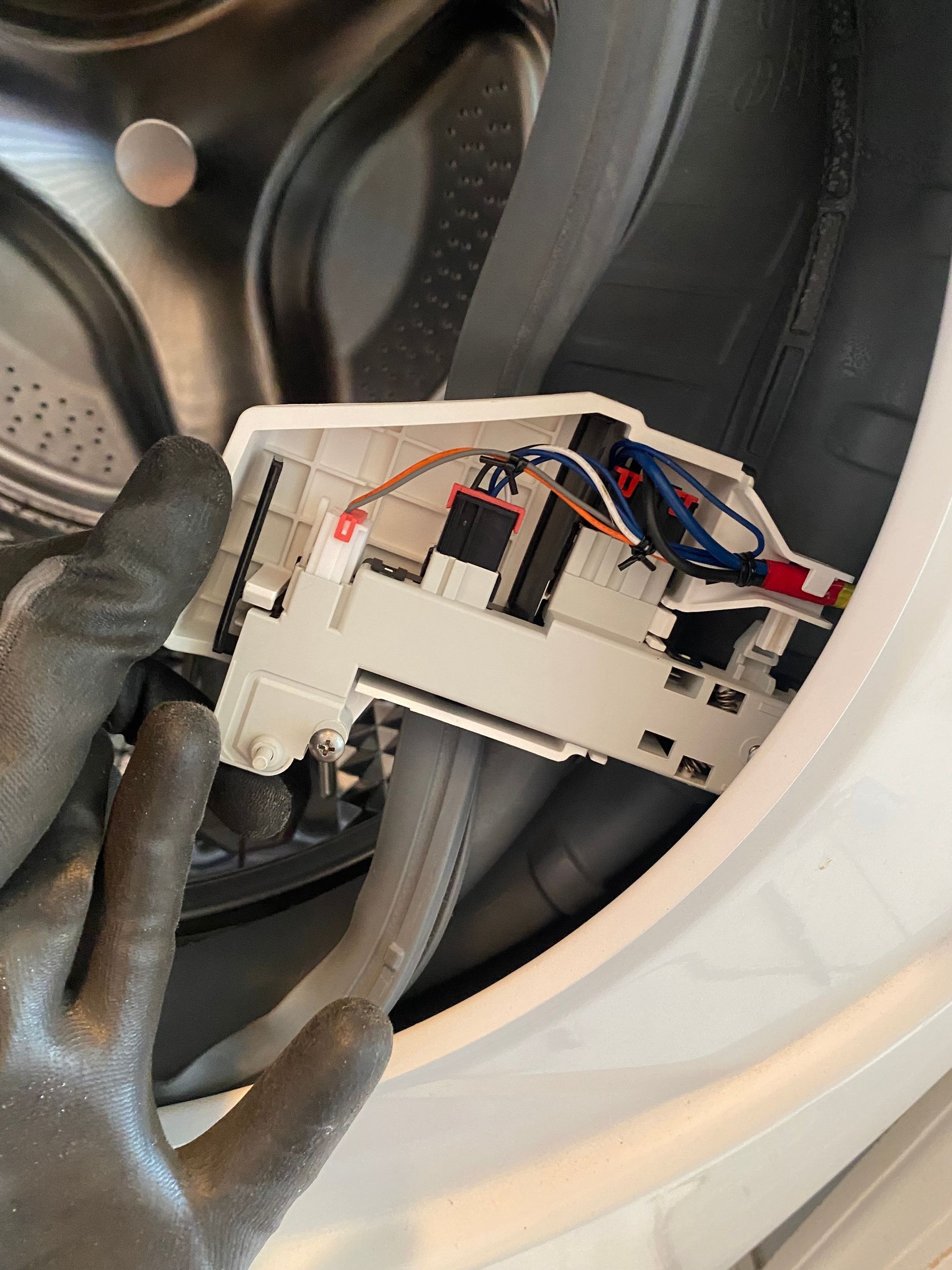 Gloved hand holding a white washing machine door lock mechanism, wires visible, next to the machine's door opening.