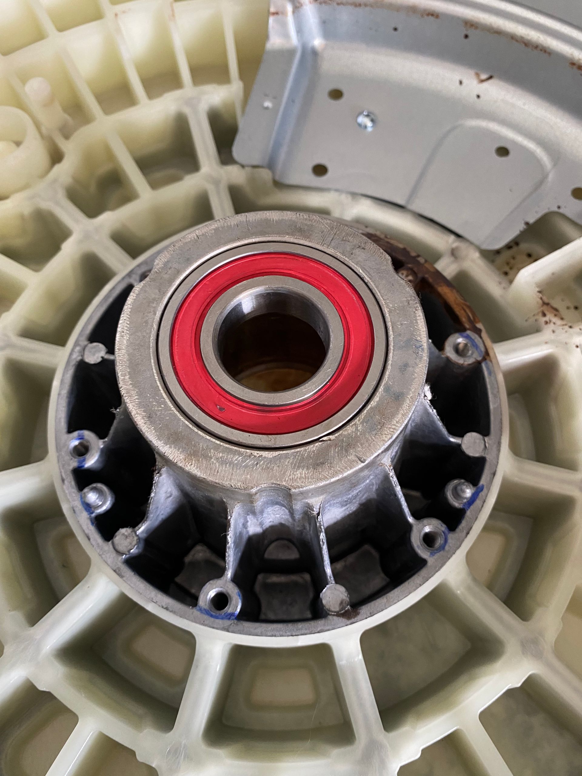 Close-up of a washing machine's inner components. A gray metal bearing with a red seal sits on a white plastic base.