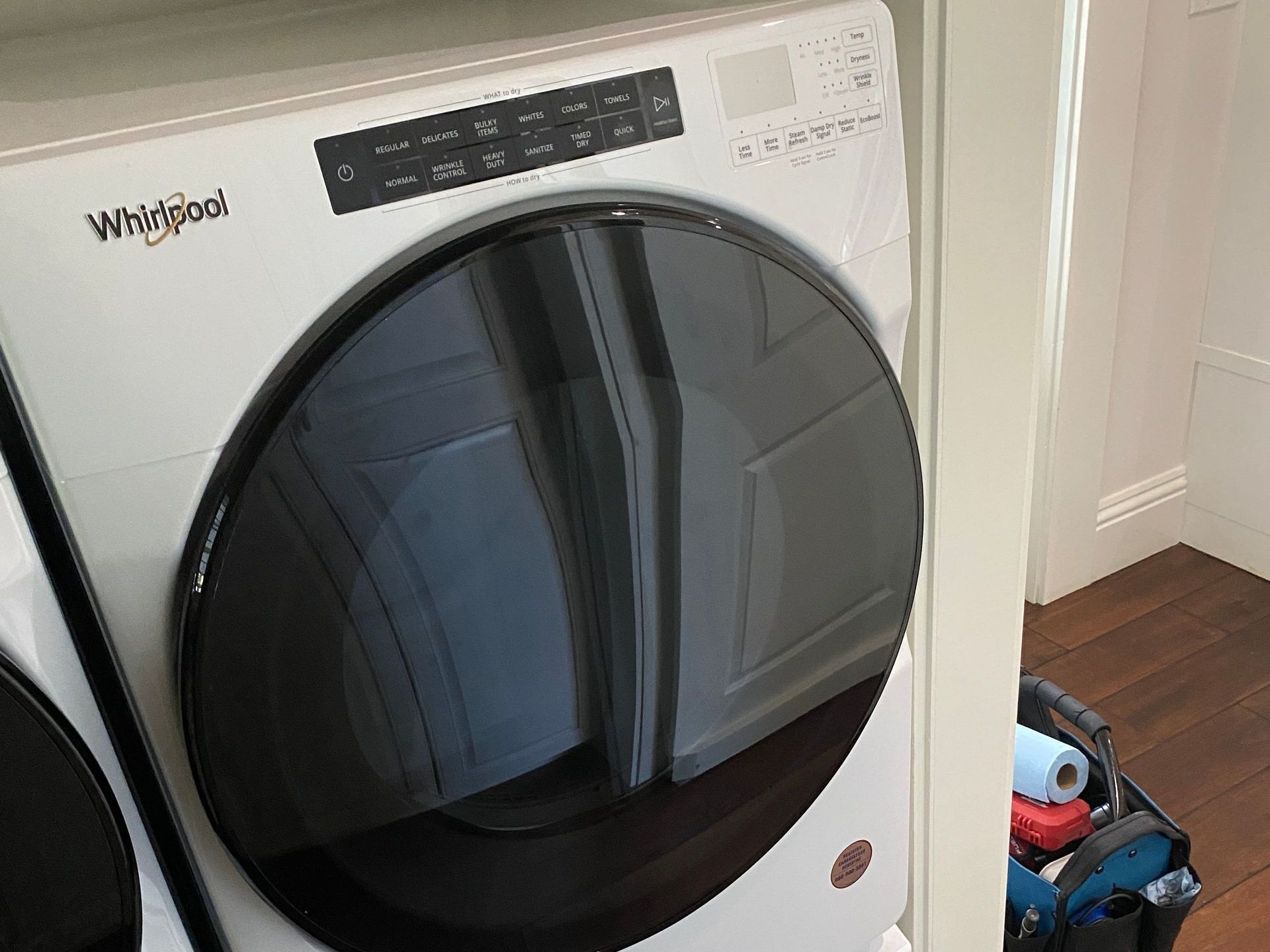 White Whirlpool washer and dryer stacked, reflecting a room; tools visible nearby.