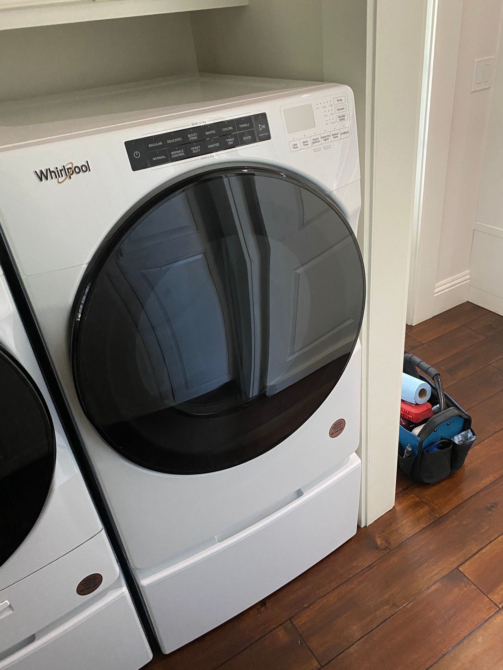 White Whirlpool dryer with a large black door, in a laundry room with a work bag visible.