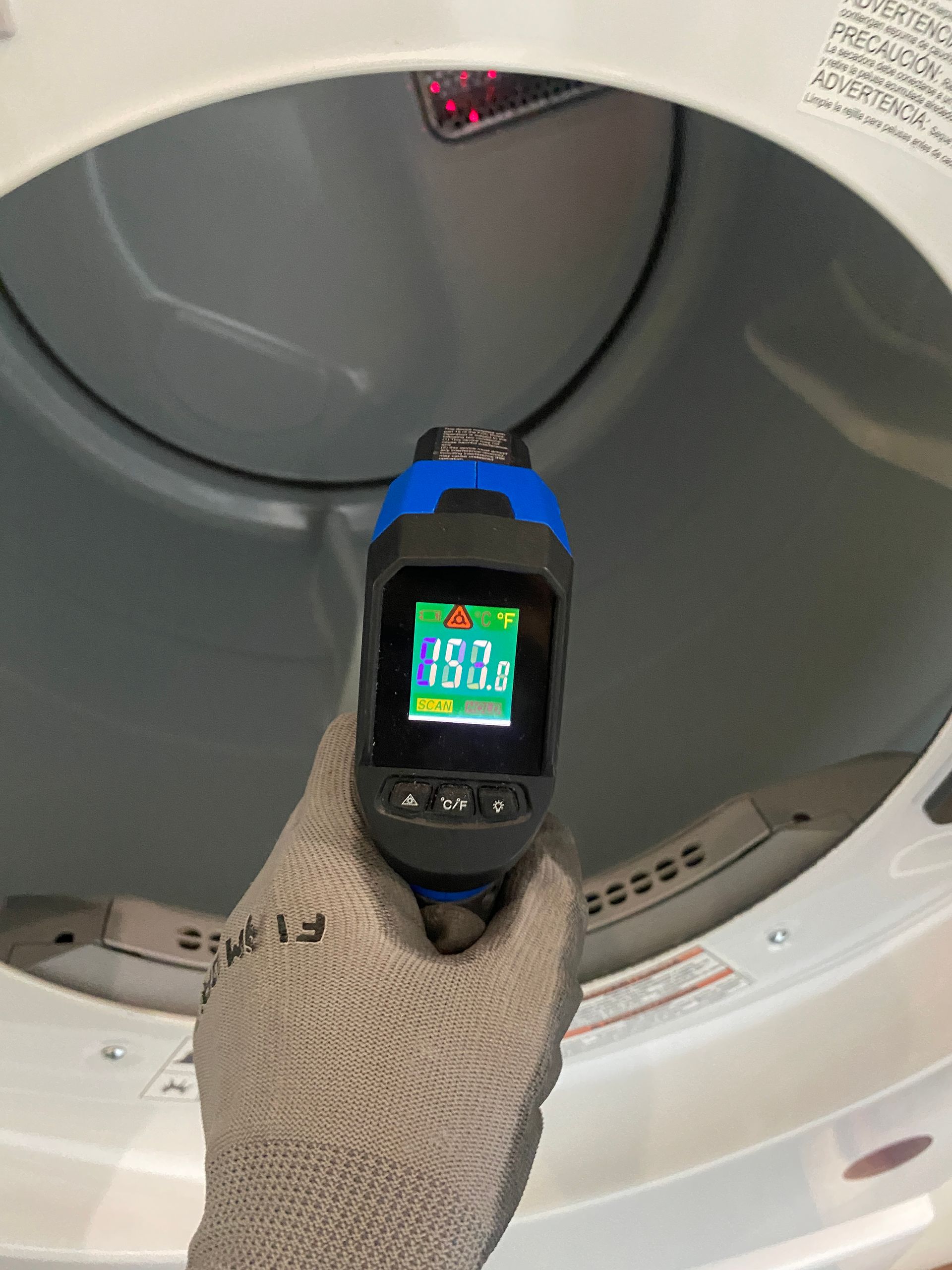 A person in a gray glove holds a temperature gun inside a white dryer, displaying 150°F on the screen.