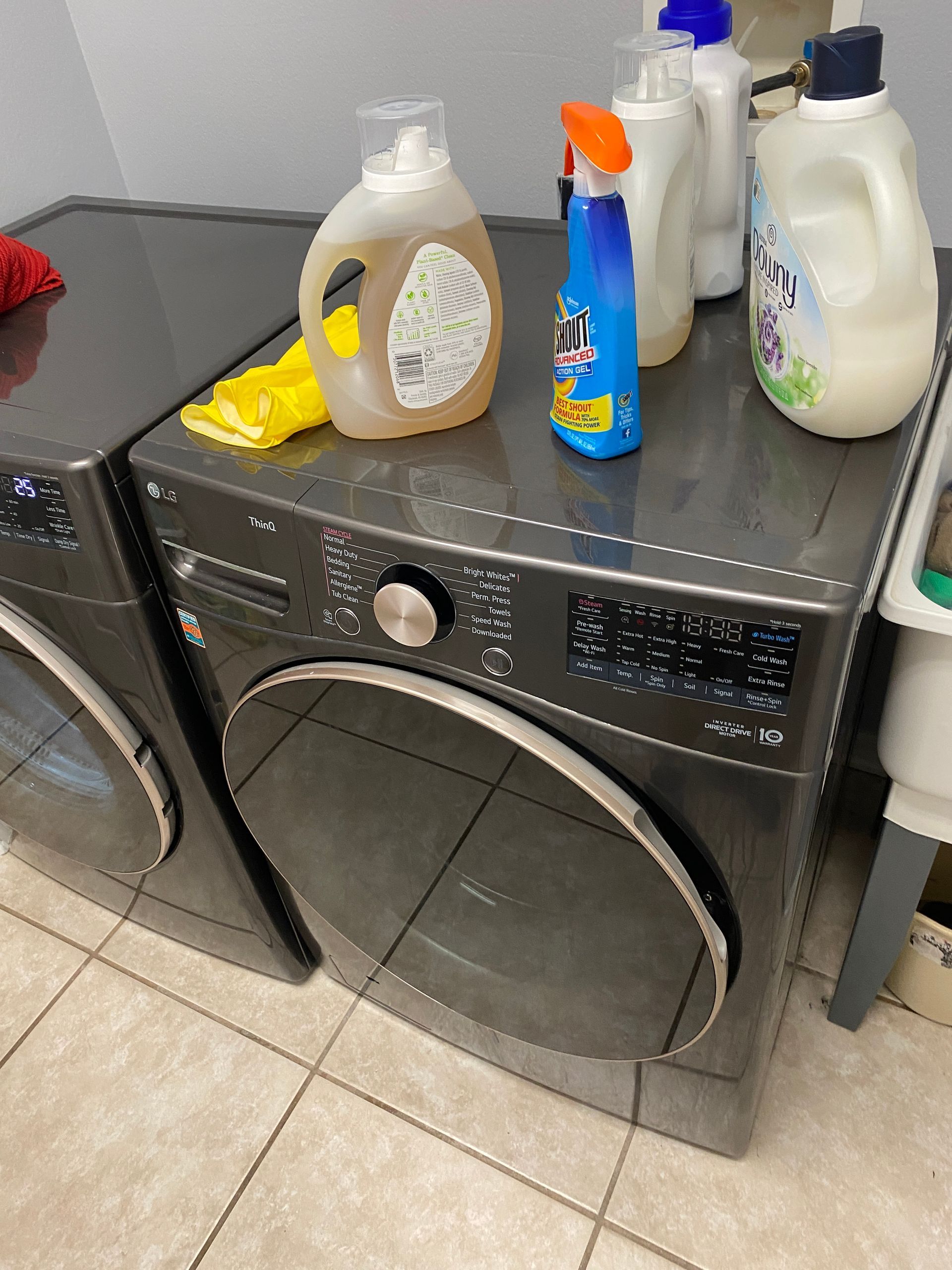 Washing machine with detergent bottles on top.  Gray appliances in a laundry room.  Yellow gloves on the left.