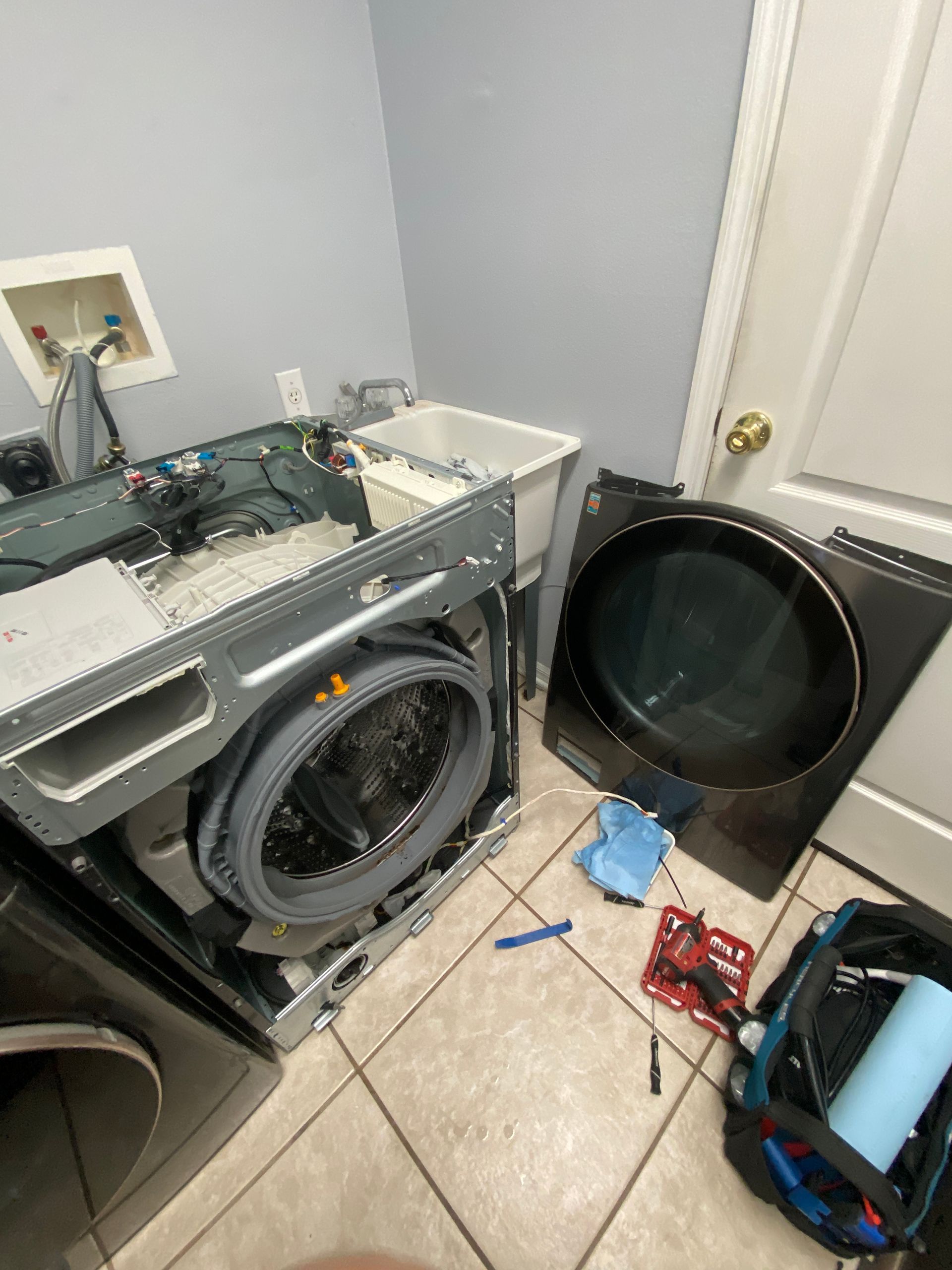 Washing machine with open front in a laundry room, parts and tools on the floor.