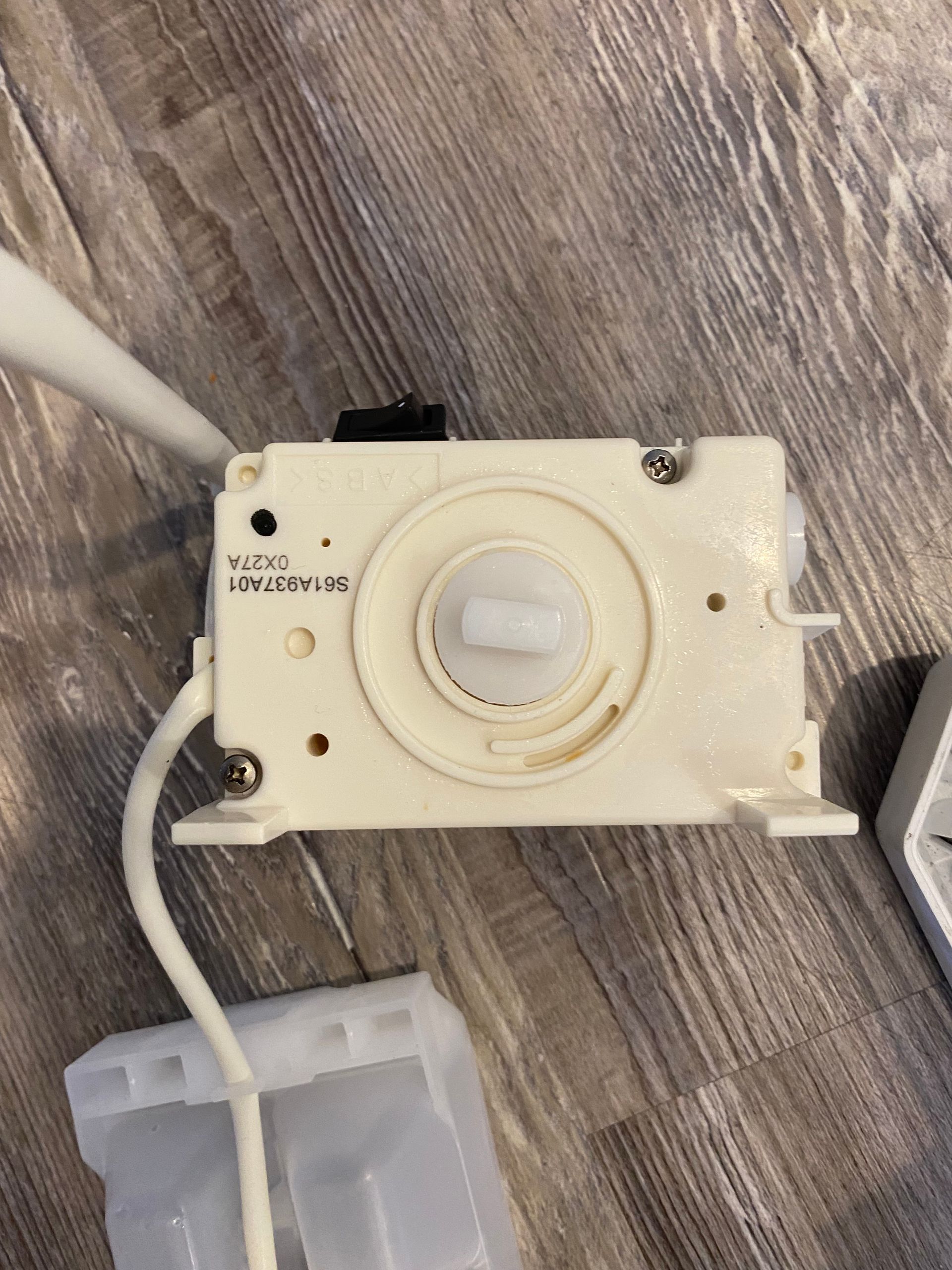 Off-white plastic appliance component with a dial and attached tubing on a wooden surface.