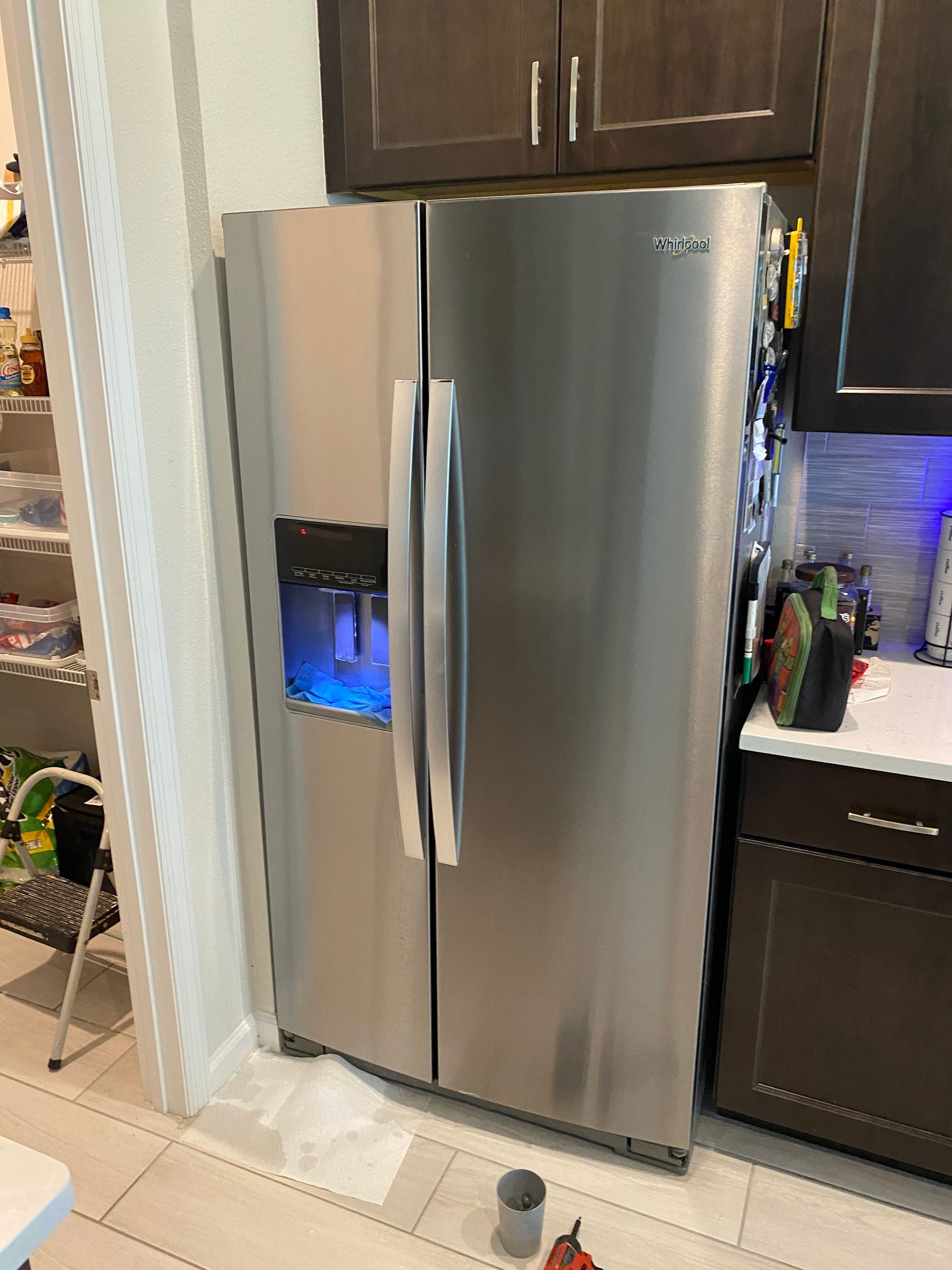 Stainless steel Whirlpool side-by-side refrigerator in a kitchen, with a water dispenser and dark cabinetry above.