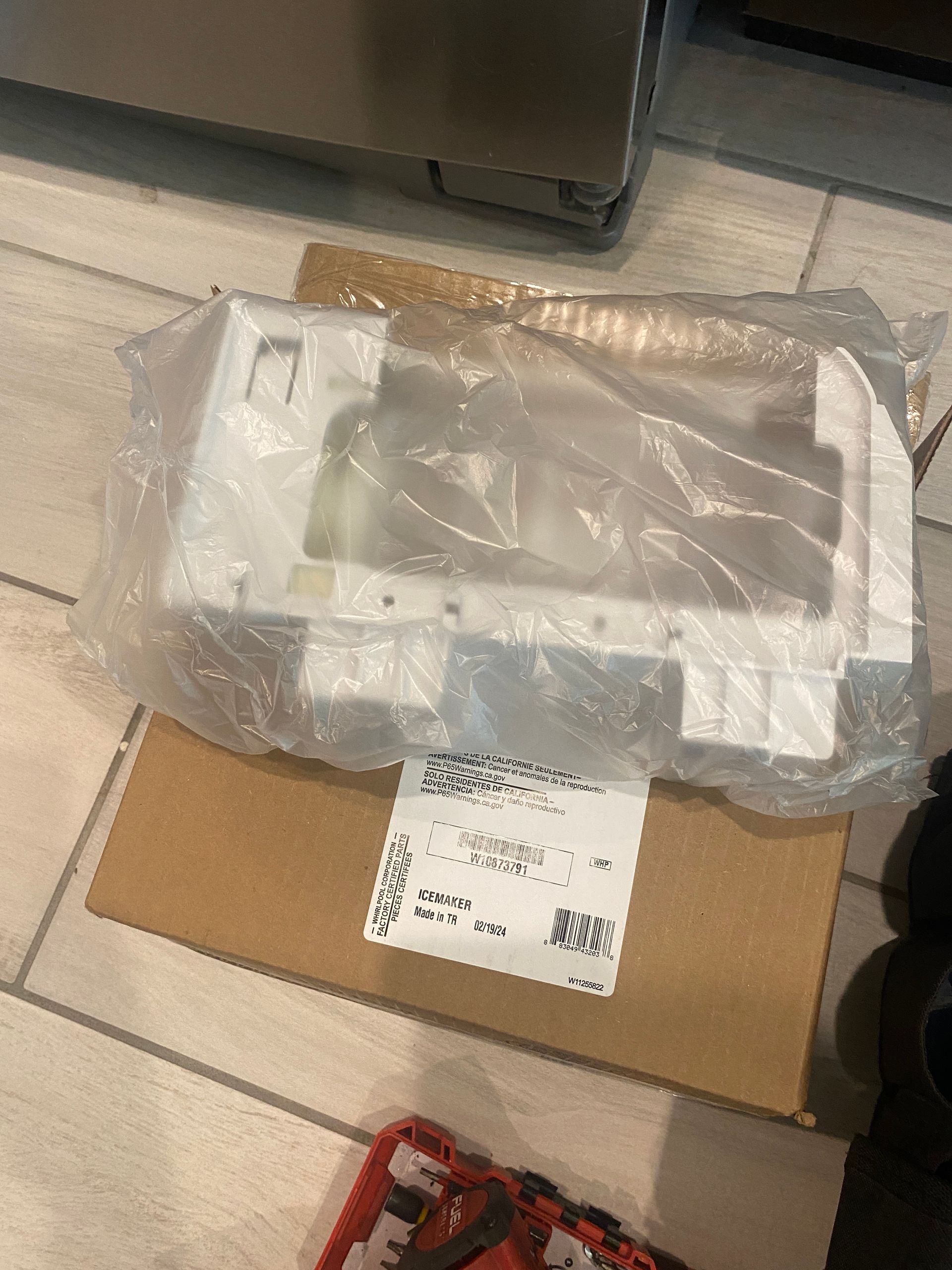 White plastic refrigerator part in a plastic bag on a cardboard box on a tiled floor.