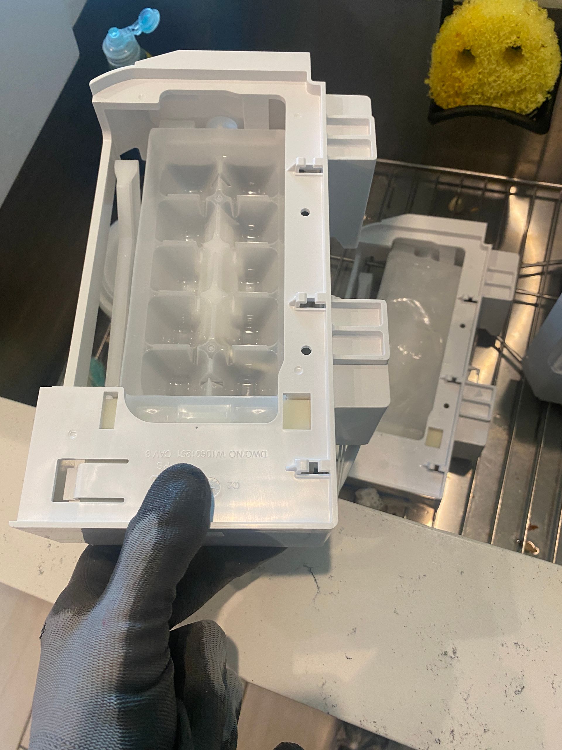 A disassembled white refrigerator ice maker held by a gloved hand.