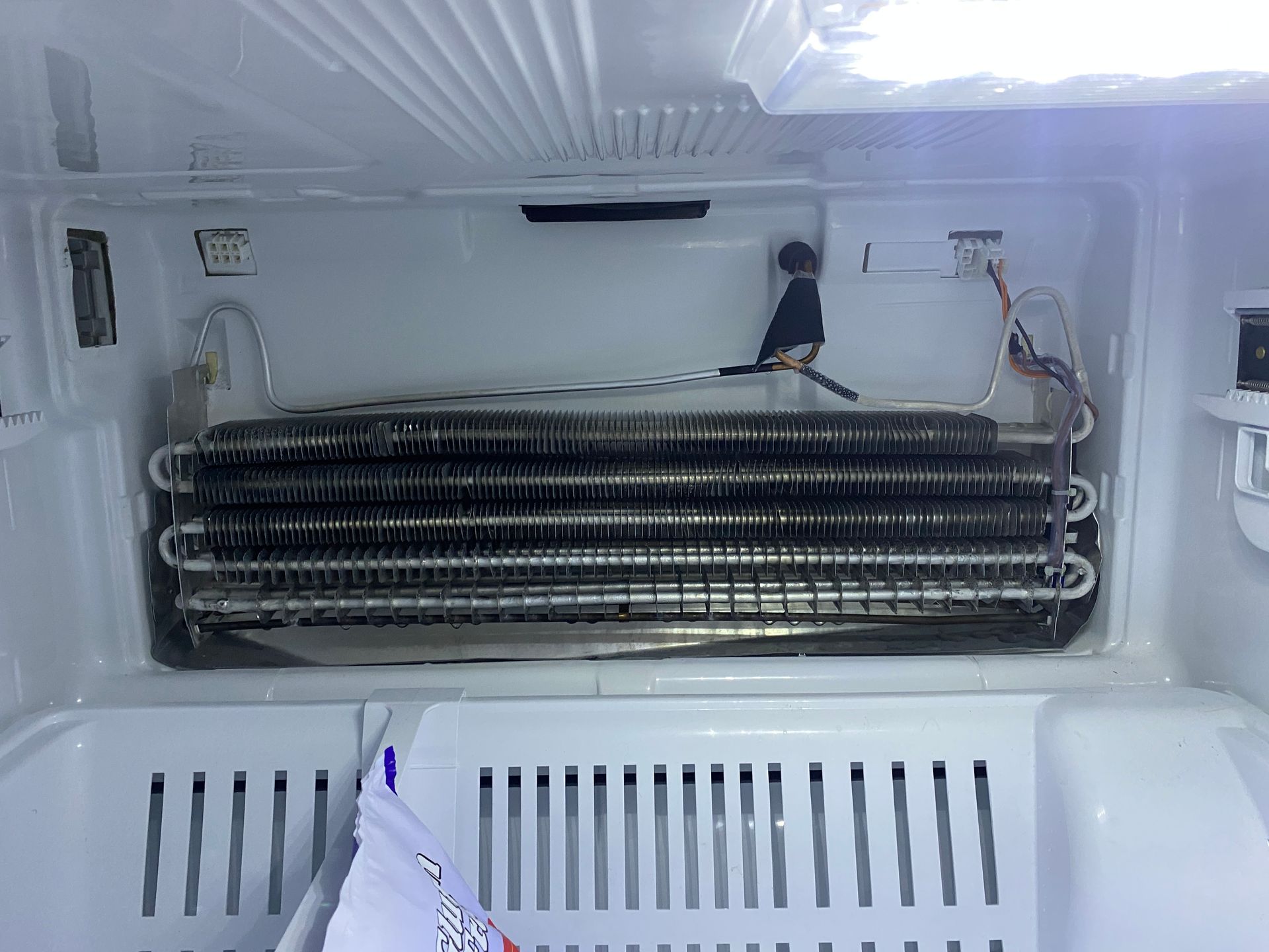 Inside of a refrigerator: frost-covered cooling coils and interior. White plastic shelving visible.