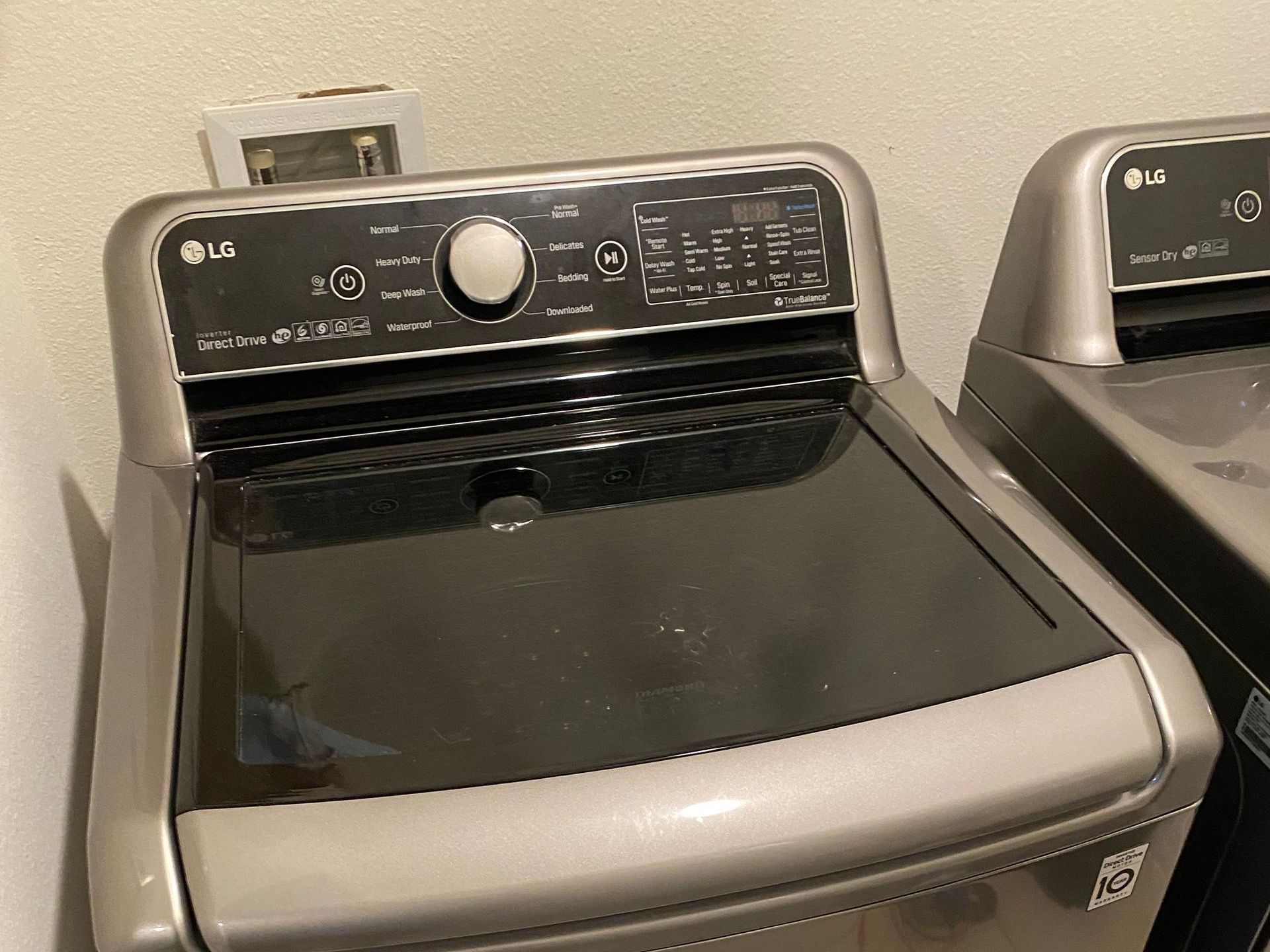 Silver LG top-load washing machine with a glass lid, indoors against a white wall.