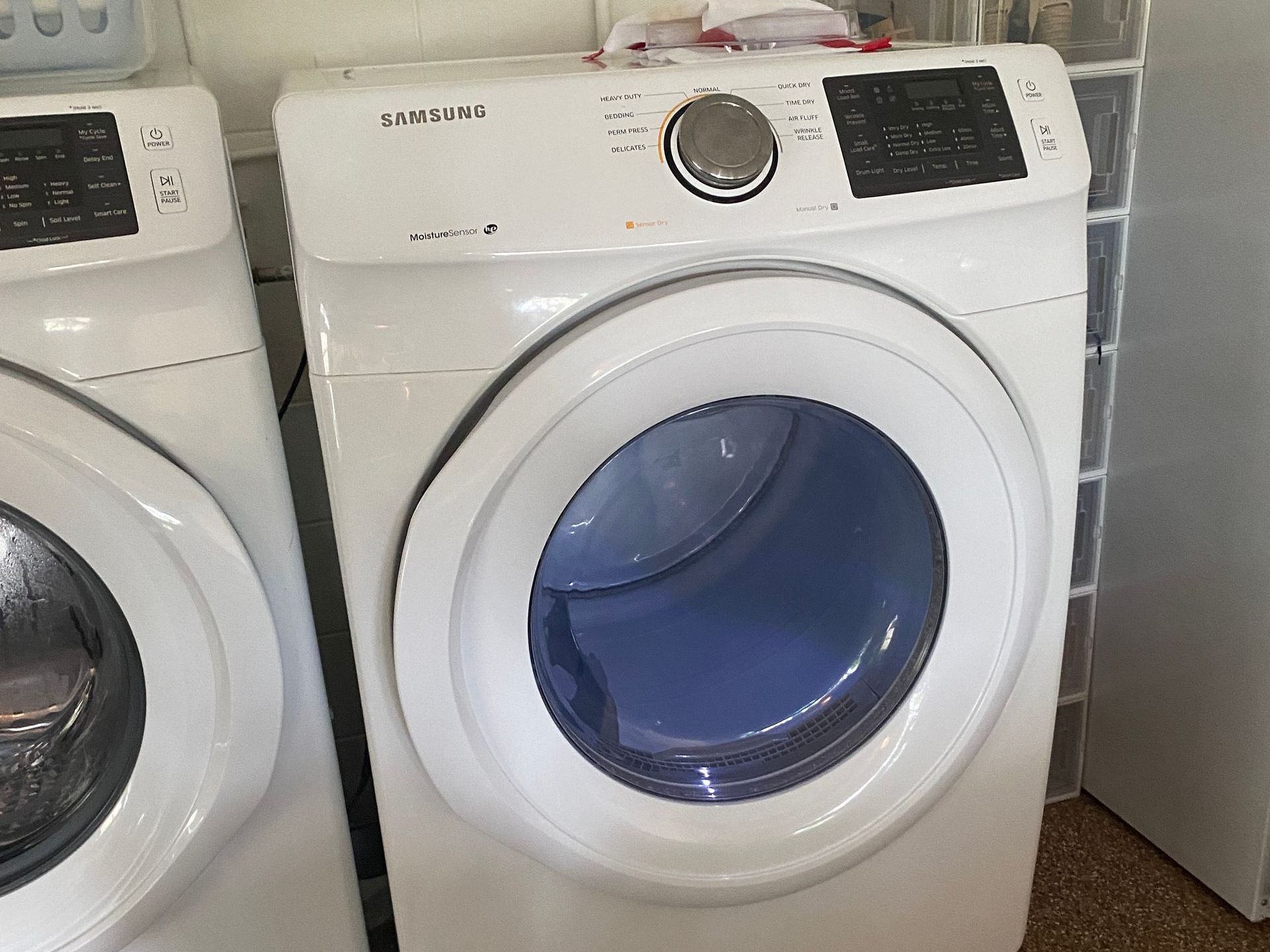 White Samsung dryer, open door, blue interior. In a laundry room setting, next to a washing machine.