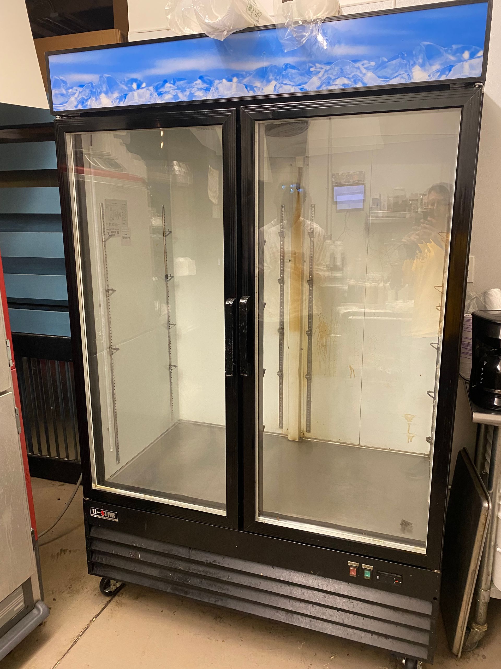 Double-door black commercial refrigerator with glass doors, illuminated top panel, and wheels, indoors.