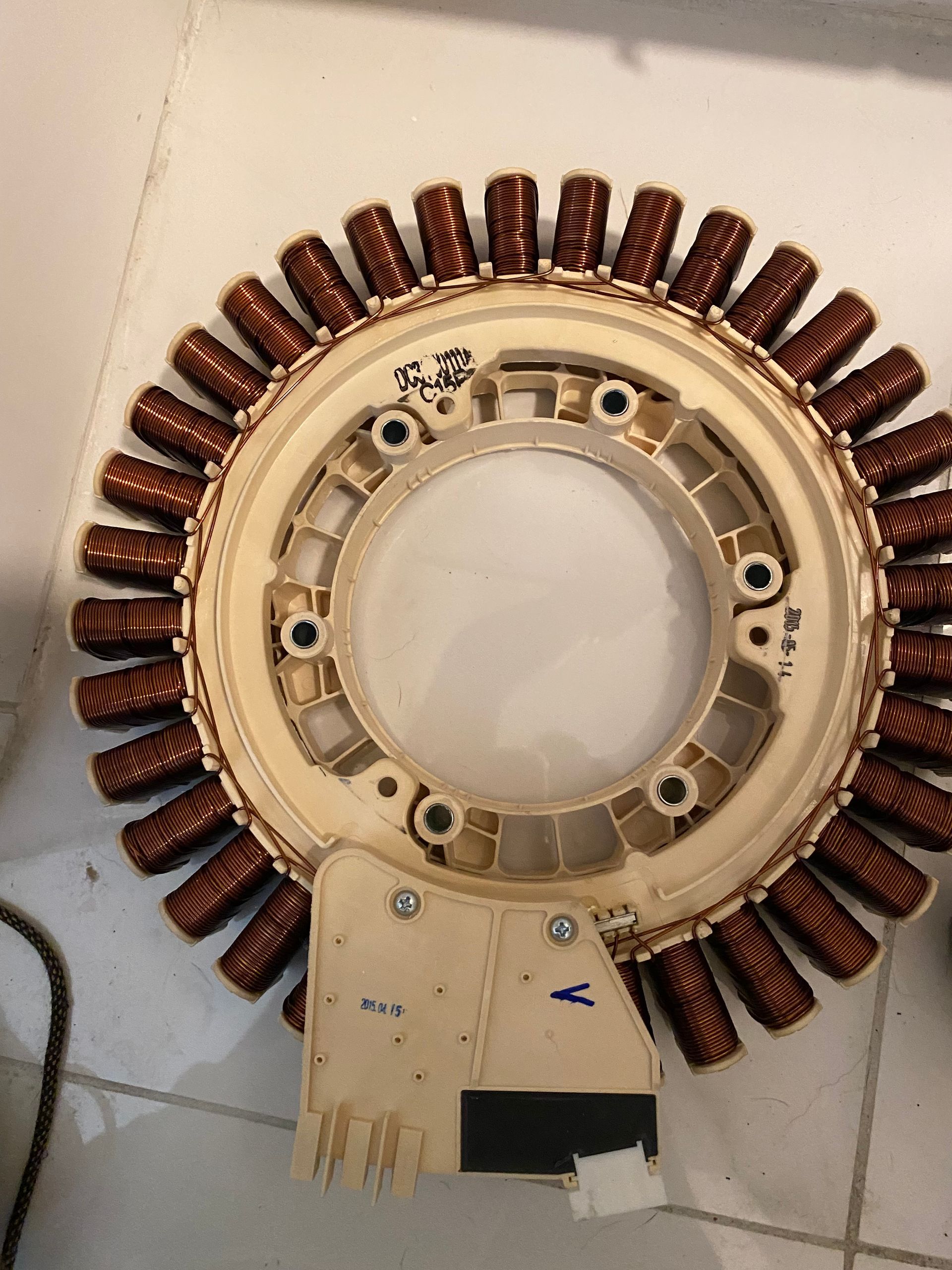 A circular electric motor stator with copper coils and a white plastic frame, set on a tiled surface.