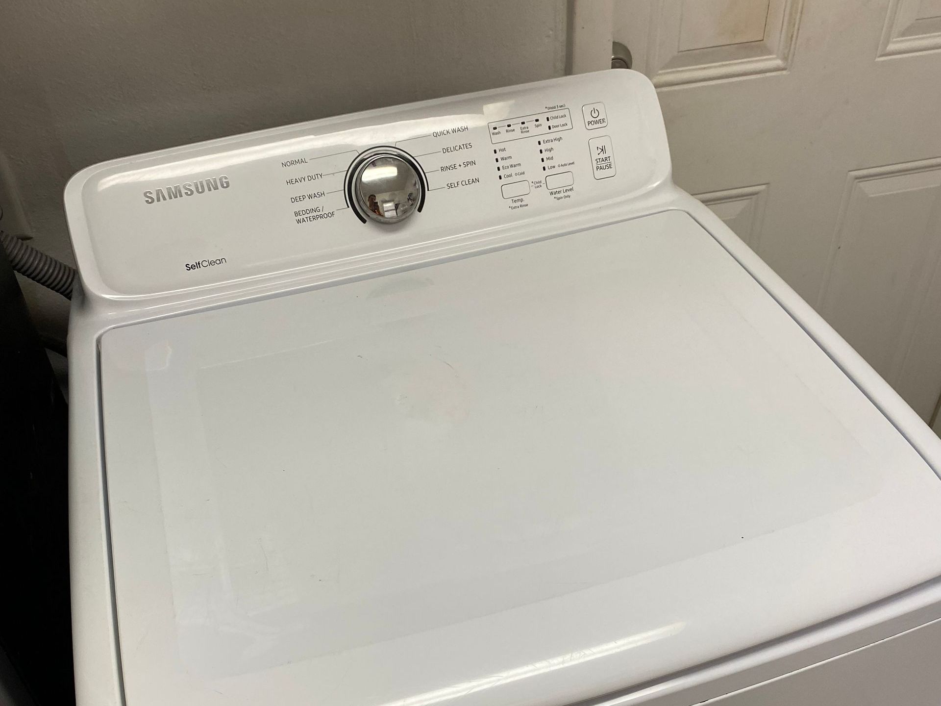 White Samsung top-load washing machine, control panel on top, in a laundry room.