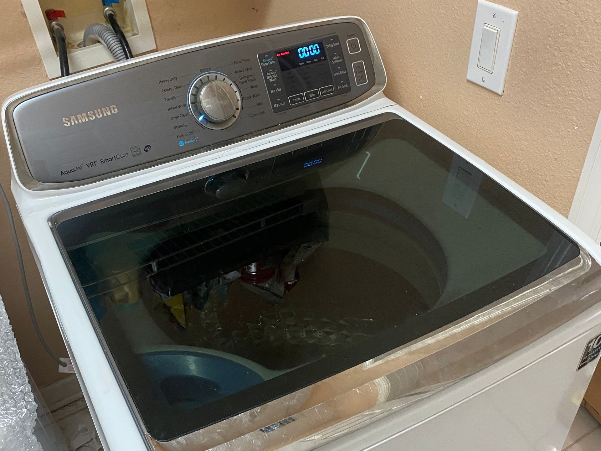 White Samsung top-load washing machine with a glass lid, against a beige wall.