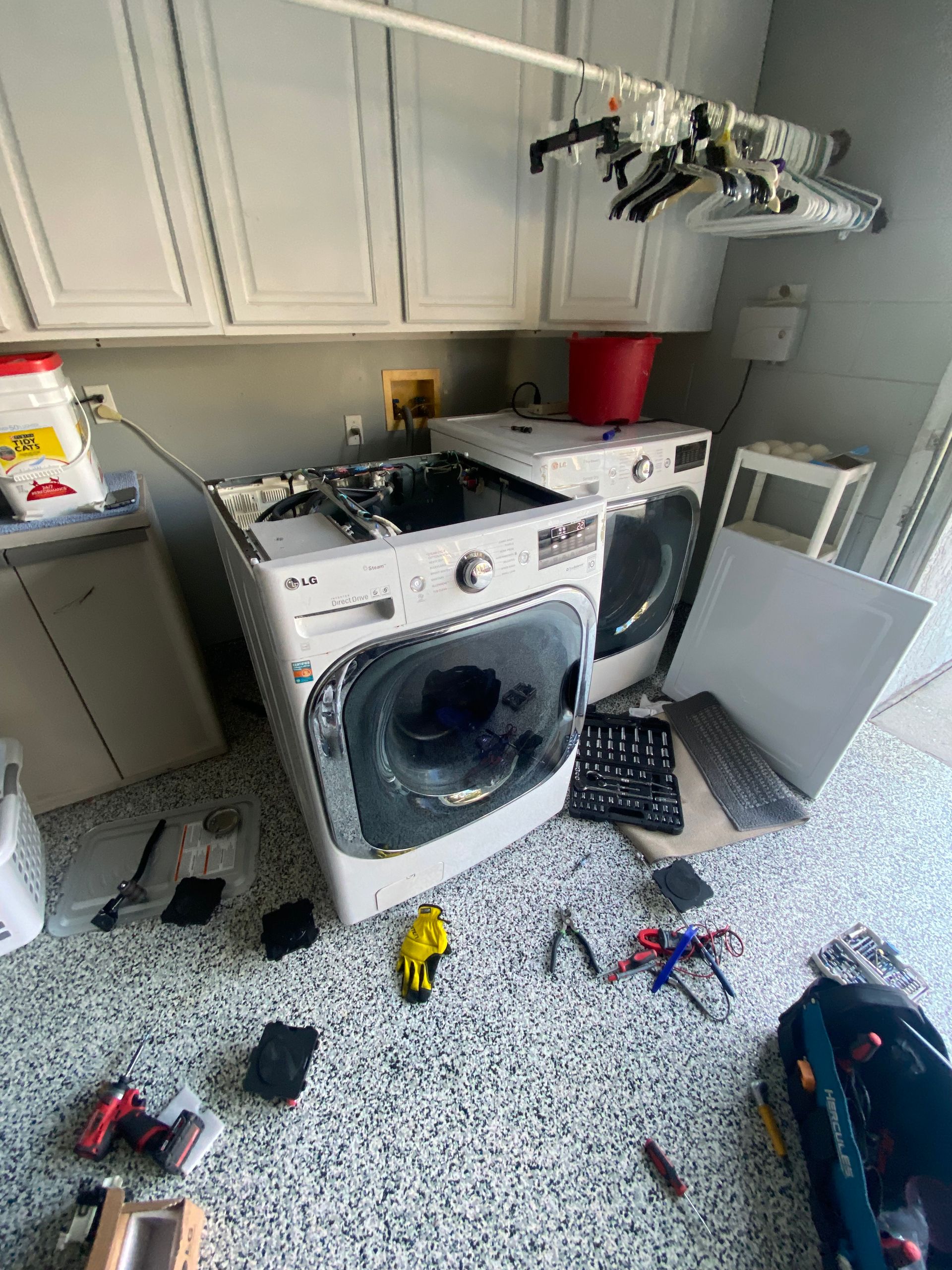 Washing machine being repaired in a laundry room. Tools and parts scattered on the floor. Cabinets above.